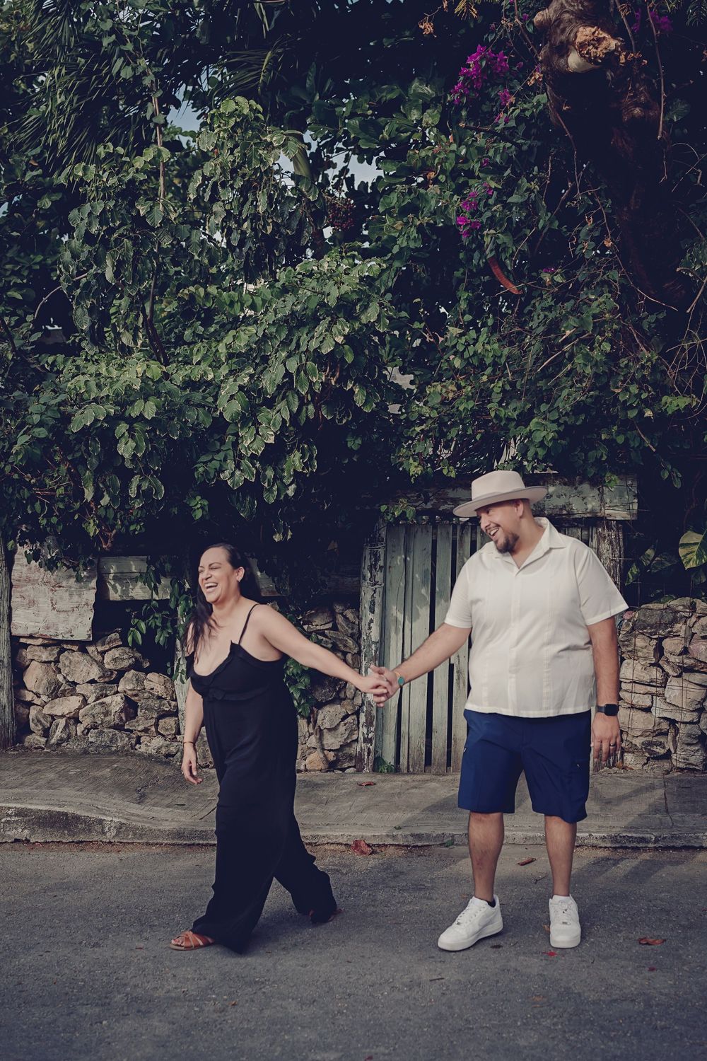 Two people sit on a low stone wall while performing playful falling or leaning poses together in an outdoor setting.