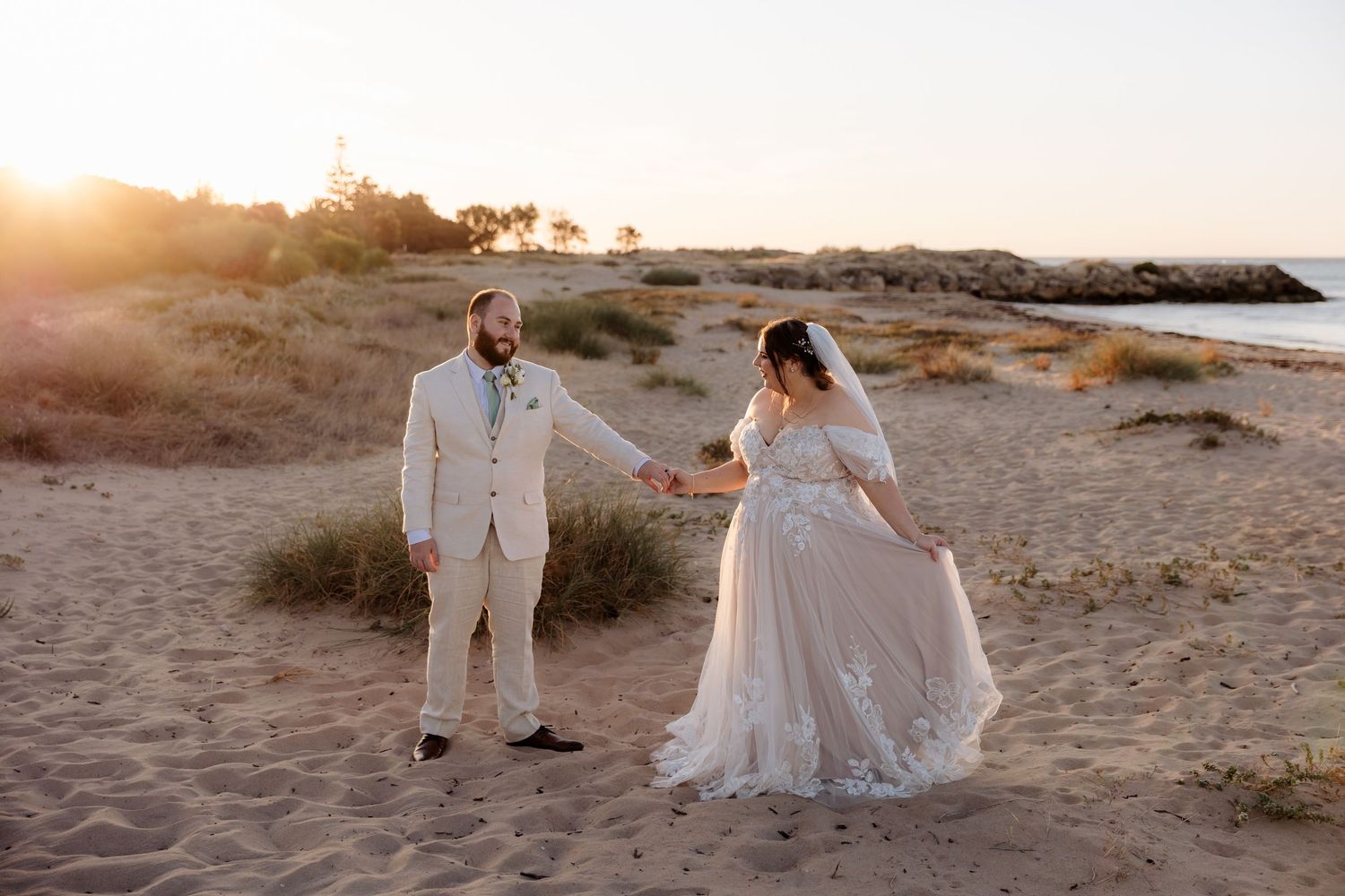 Two people in wedding attire walk along a coastal beach at sunset as golden light illuminates the rocky shoreline.