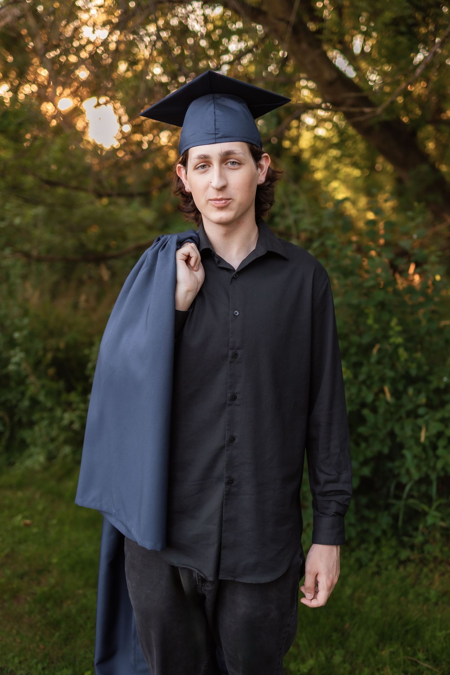 Graduate in cap and black button-down shirt stands outdoors with trees and warm sunlight in background. Novi Michigan Senior Photographer