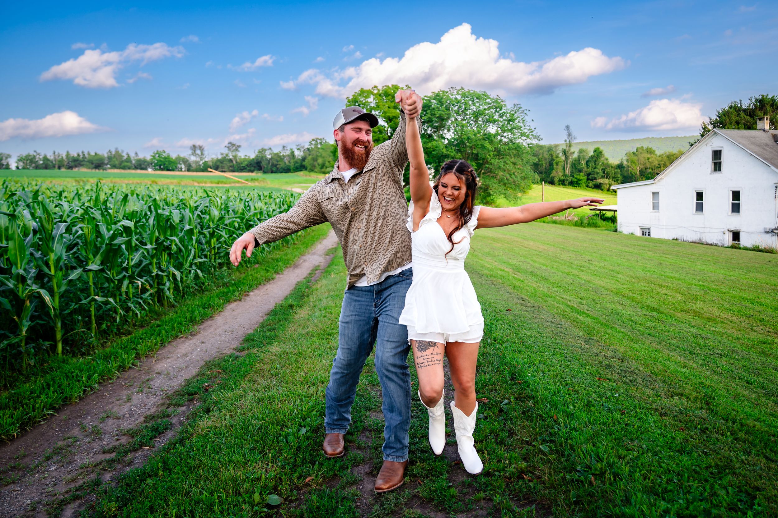 A sunny rural landscape featuring a dirt path between green cornfields and farmland.