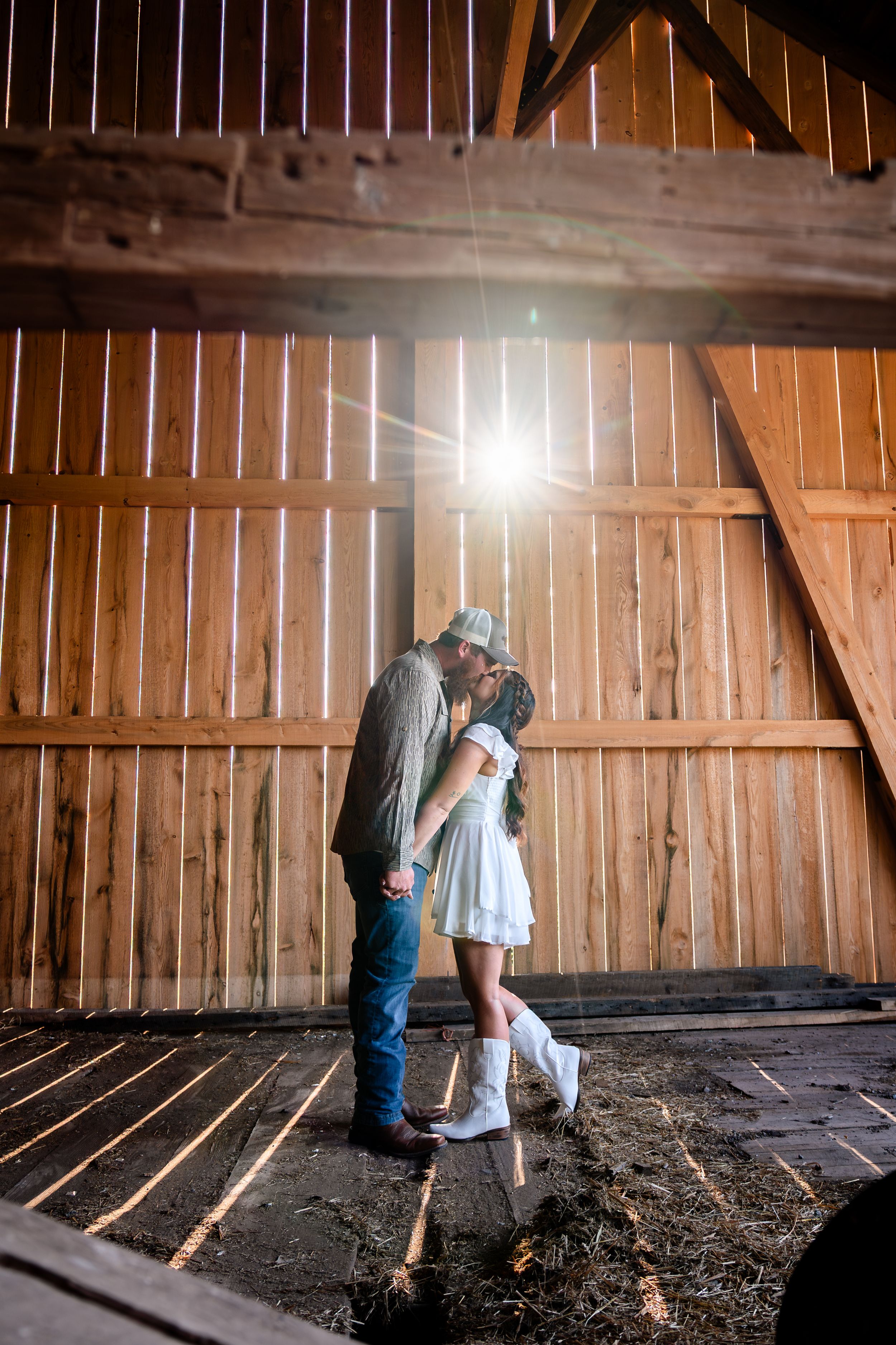 A couple shares an intimate moment inside a rustic wooden barn with sunlight streaming through.