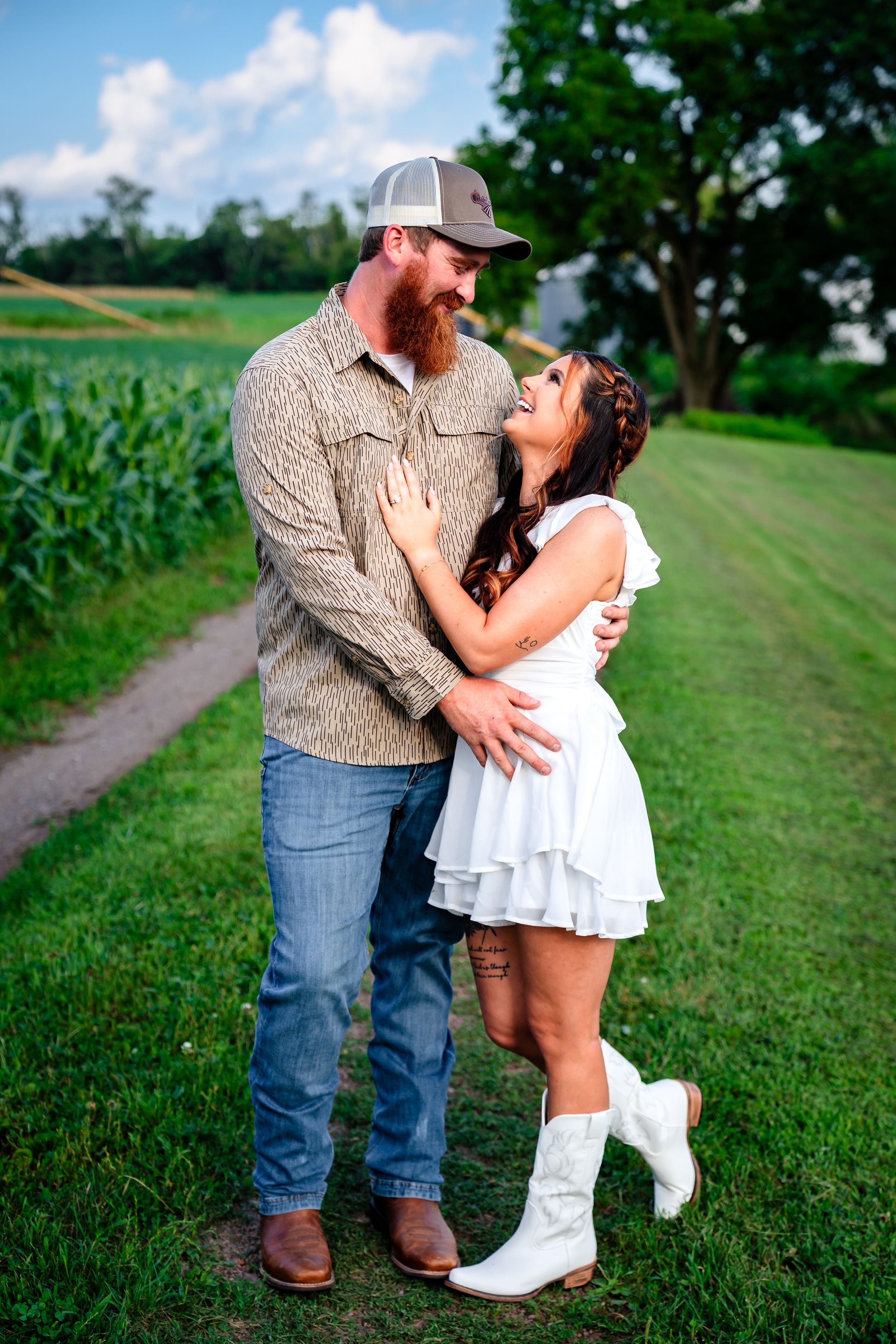 A couple in casual attire shares a romantic moment on a grassy path near a cornfield.