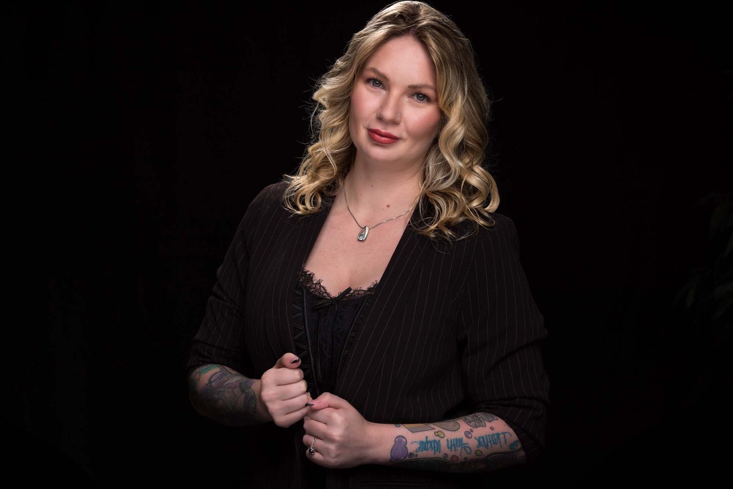 Professional studio portrait with black background featuring someone in black attire with blonde wavy hair and visible tattoos.