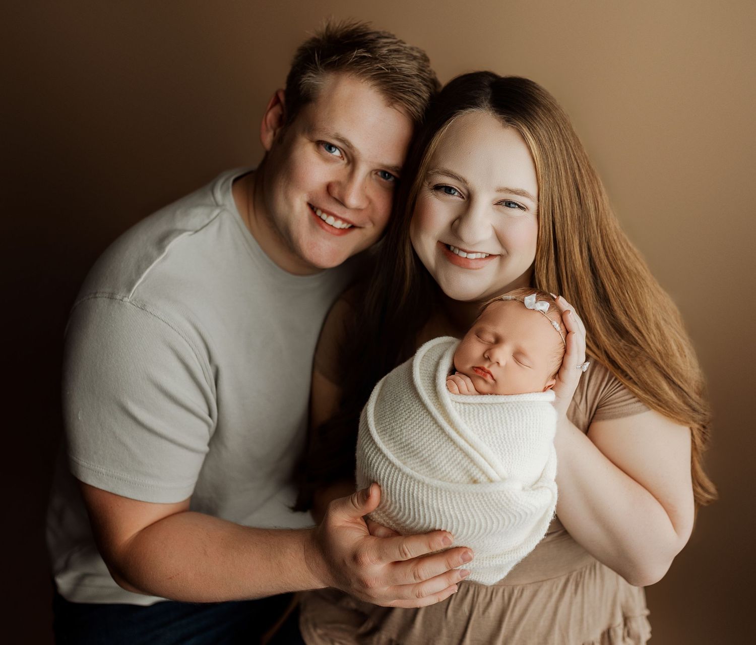 Smiling couple poses with their swaddled newborn baby for a professional studio portrait photo.