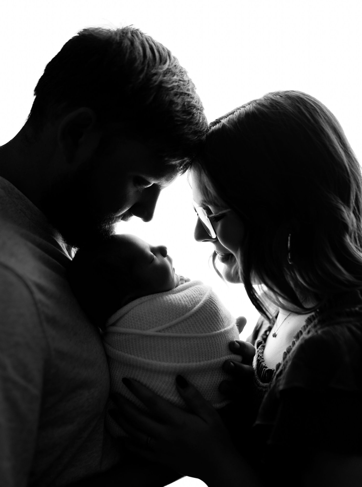 Black and white silhouette of parents lovingly cradling their newborn baby against a bright white background.