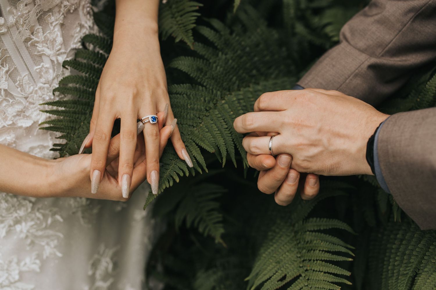Close up view of hands showing engagement rings with lush green fern leaves in the background.