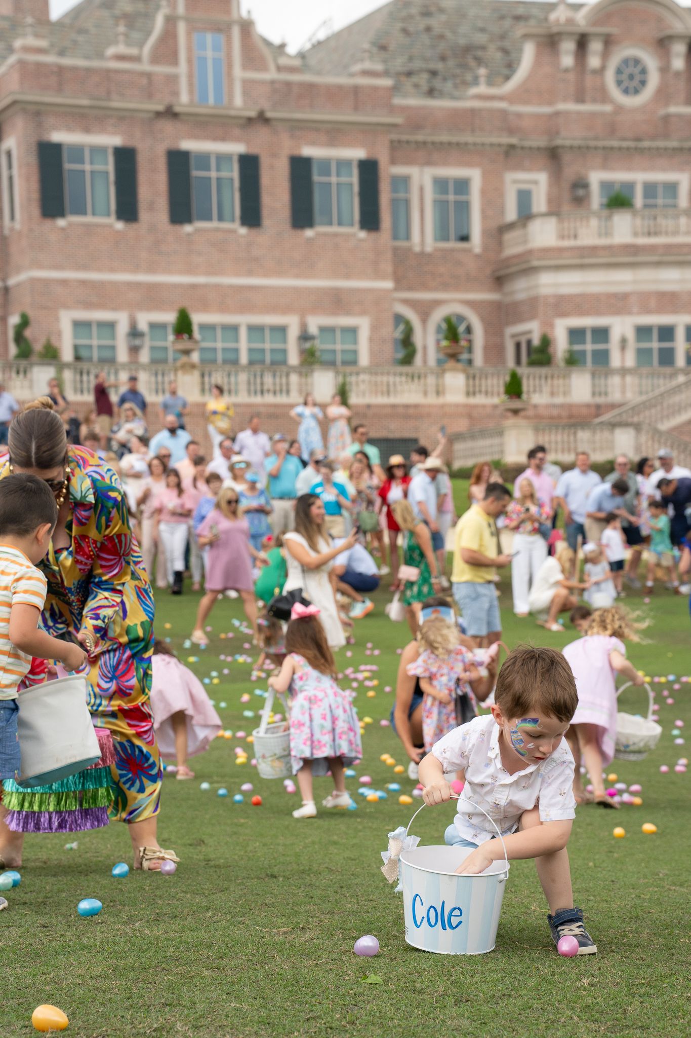 Children gather on a lawn for an Easter egg hunt event at a grand brick mansion, collecting eggs in baskets and buckets.