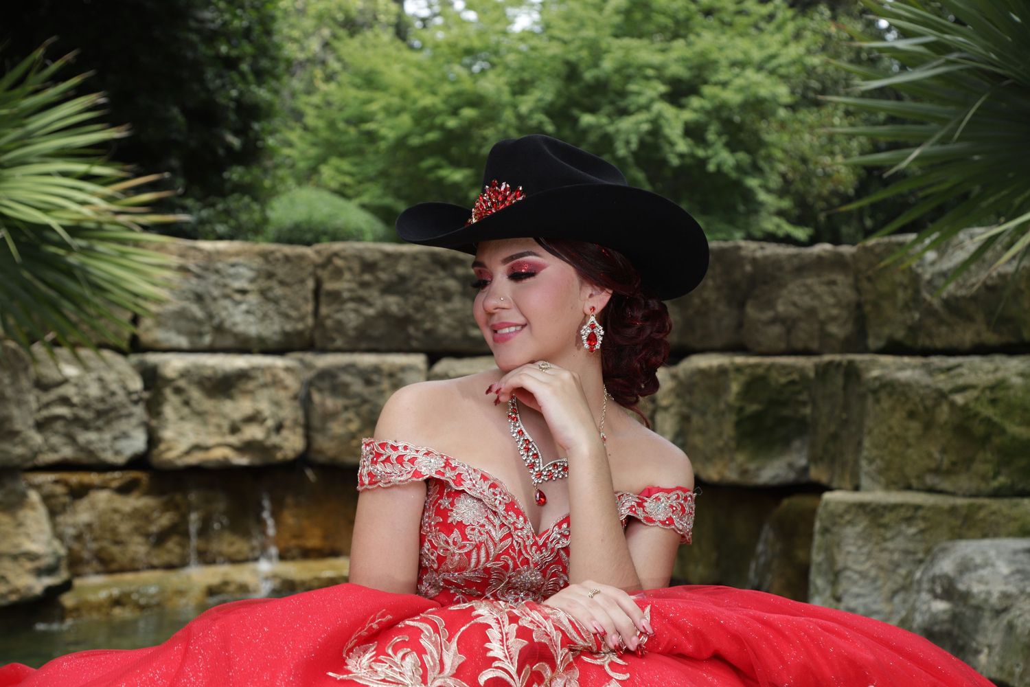 A person in a vibrant red quinceañera dress poses gracefully in a lush garden setting for outdoor portraits.