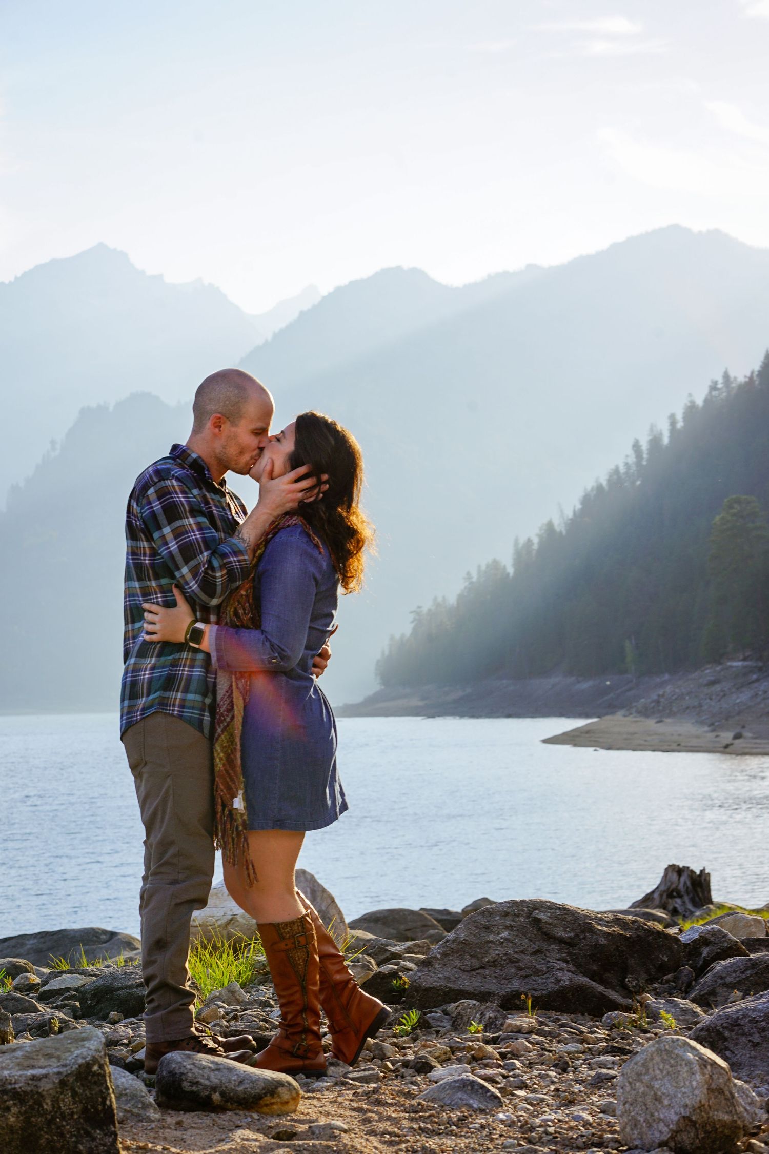 A couple shares a romantic moment on rocky terrain overlooking a misty mountain lake at sunset.