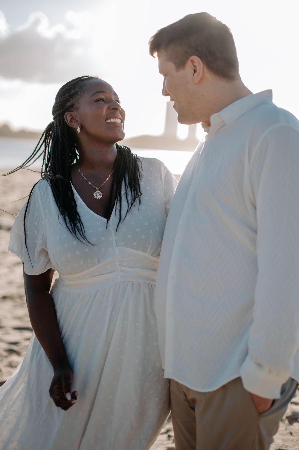A couple shares a romantic moment on the beach at sunset wearing white outfits.
