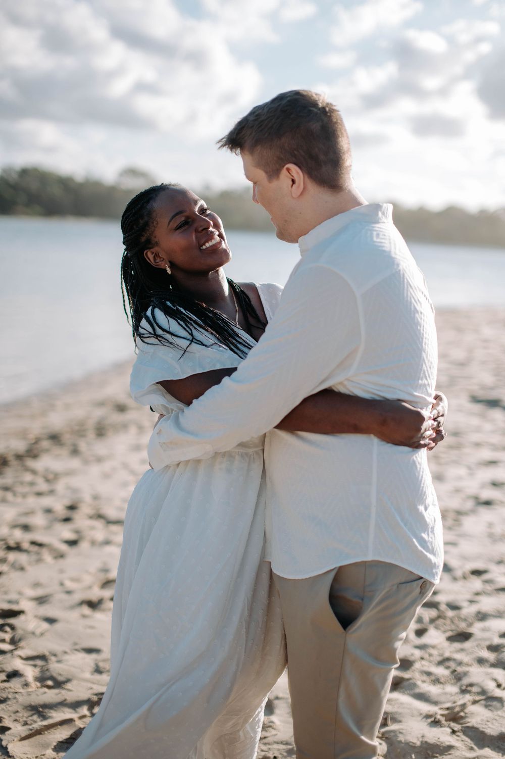 Happy couple embraces on a sandy beach at sunset wearing white clothing and sharing a joyful moment.