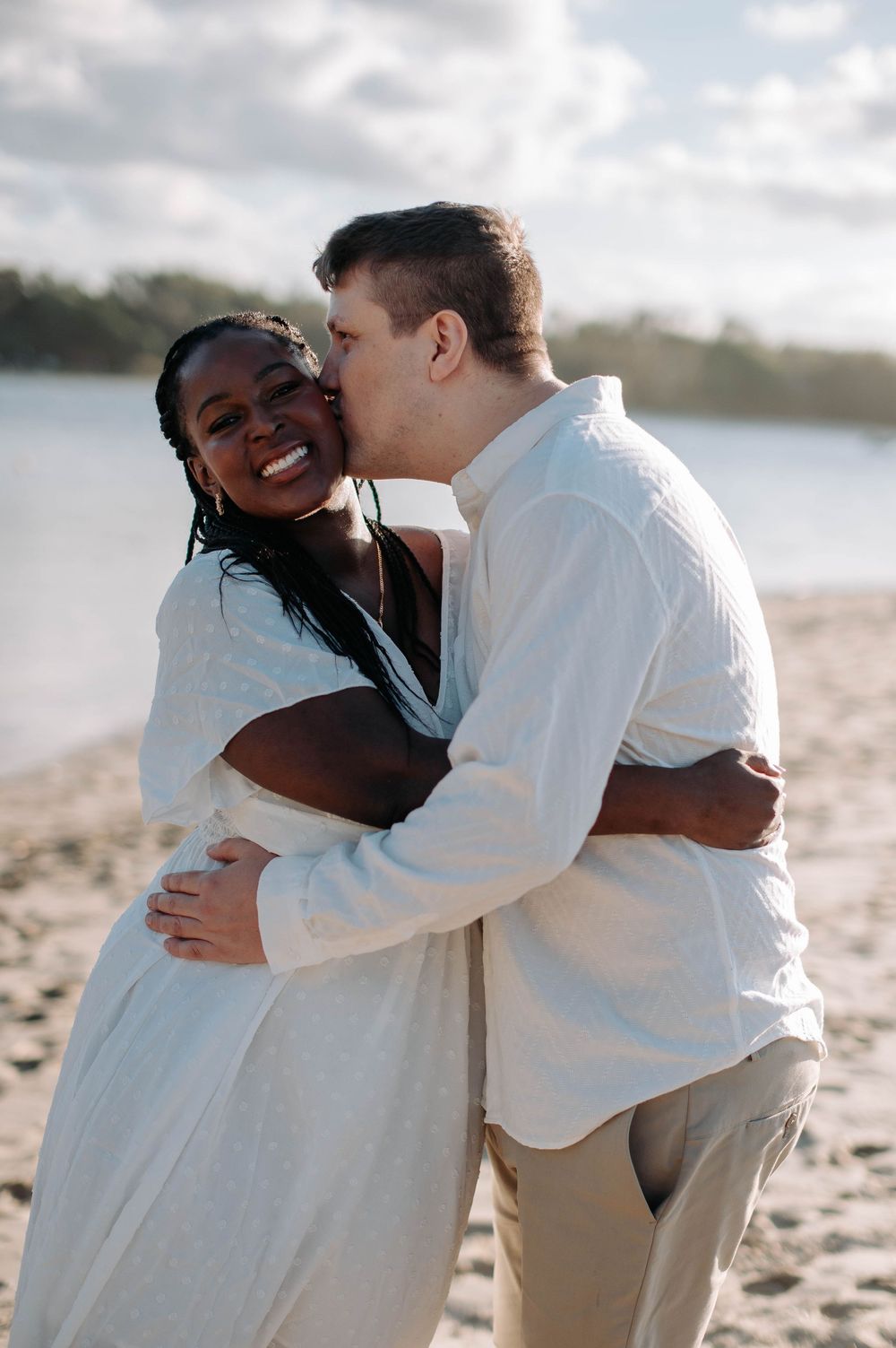A loving couple embraces and shares a joyful moment together on a sunny beach with water in the background.