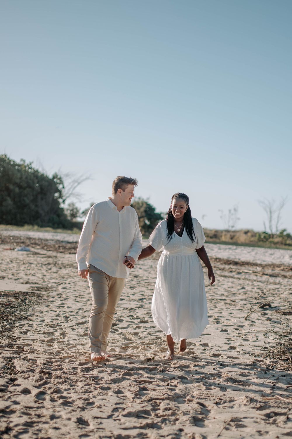 A couple walks hand in hand on a sandy beach wearing light colored clothing on a sunny day.