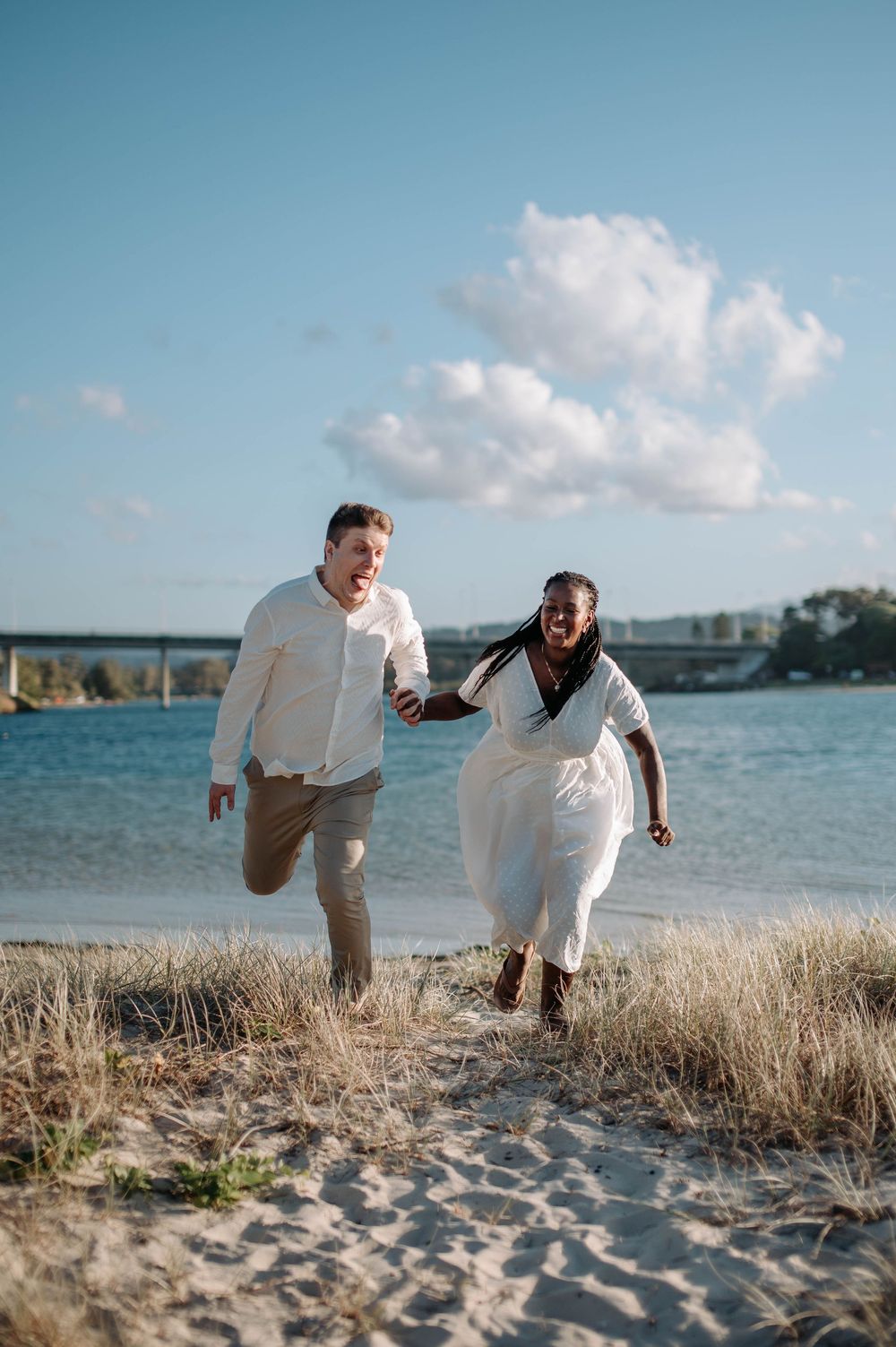 A joyful couple runs together along the beach with bright blue water and fluffy clouds in the background.
