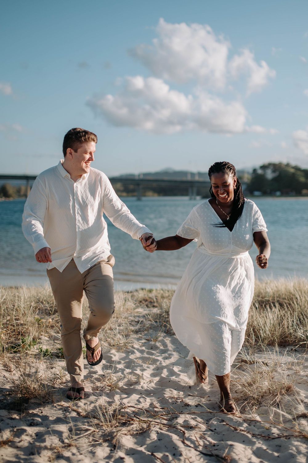 A couple in white clothing running joyfully together on a sandy beach with blue sky and water in the background.
