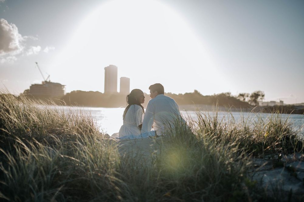 A silhouetted couple sits together by a waterfront at sunset as tall grass sways in the foreground.