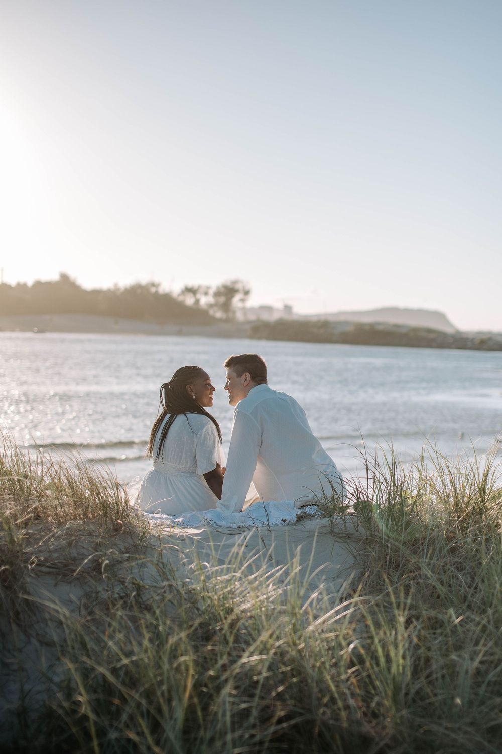 A romantic couple sits together on a beach dune at sunset, overlooking the calm waters and distant coastline.