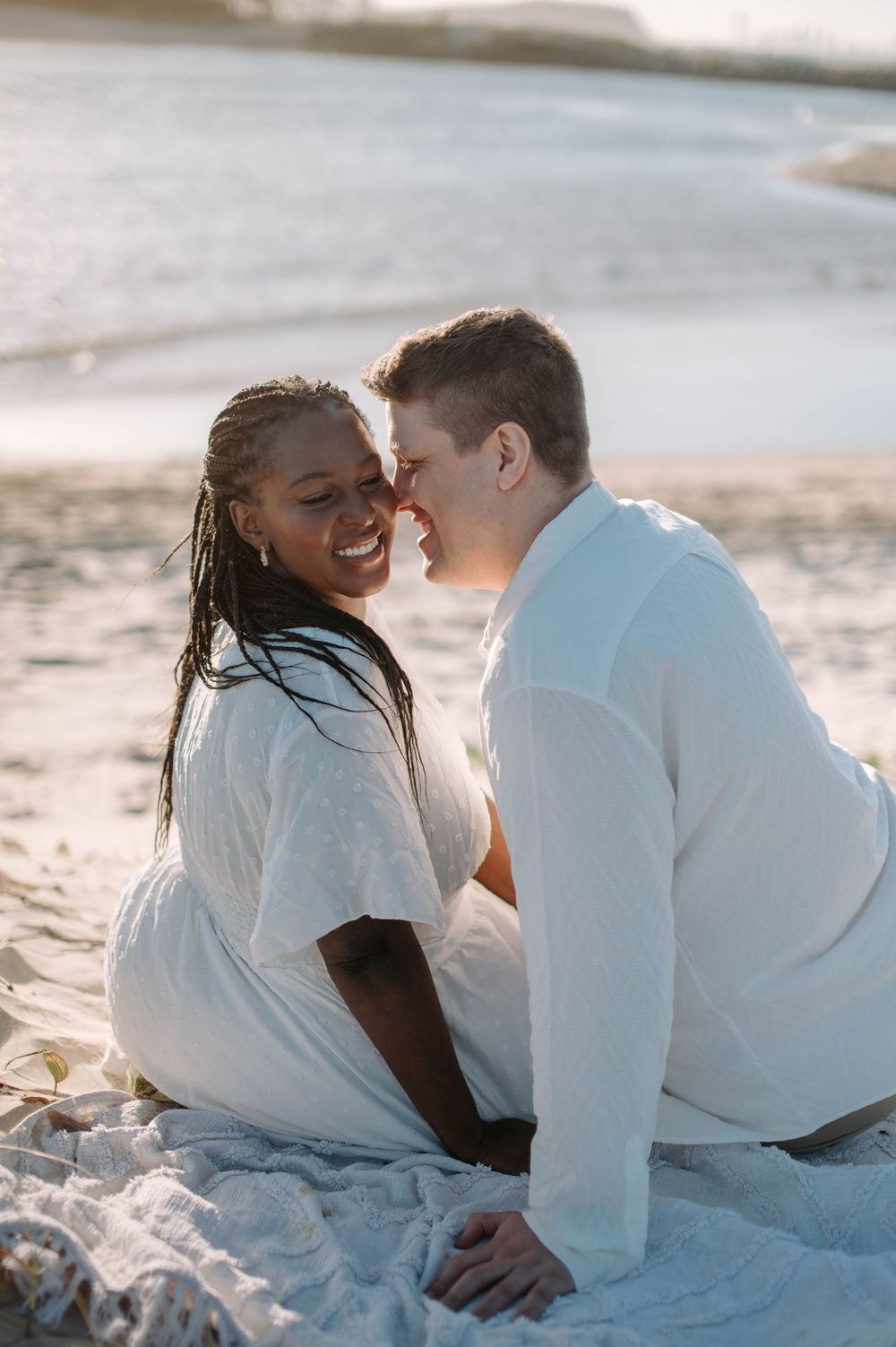 A romantic couple wearing white sits together on a sunny beach during an intimate moment at sunset.