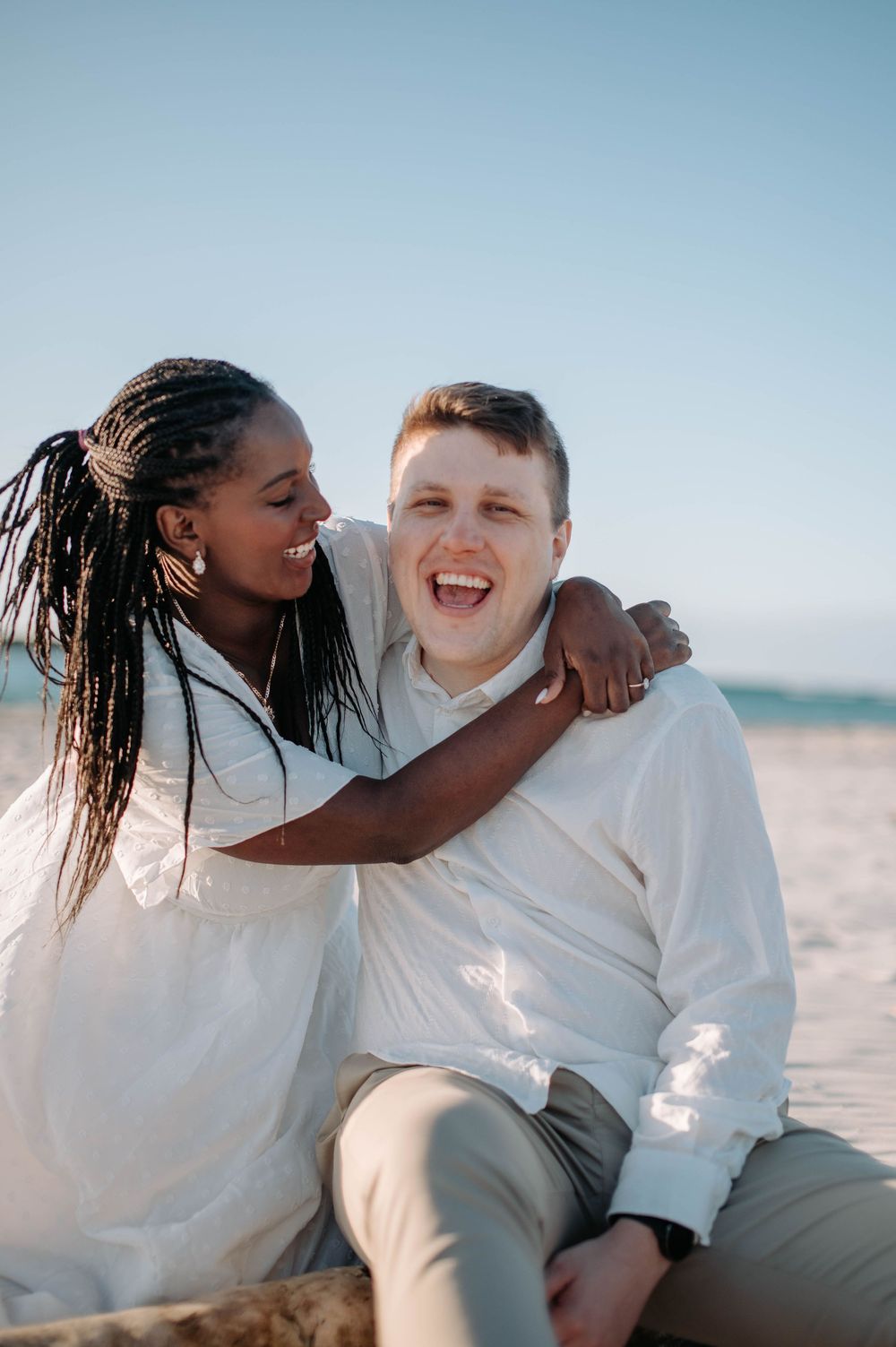 A joyful couple in white outfits share a romantic moment together on the beach during a sunny afternoon.