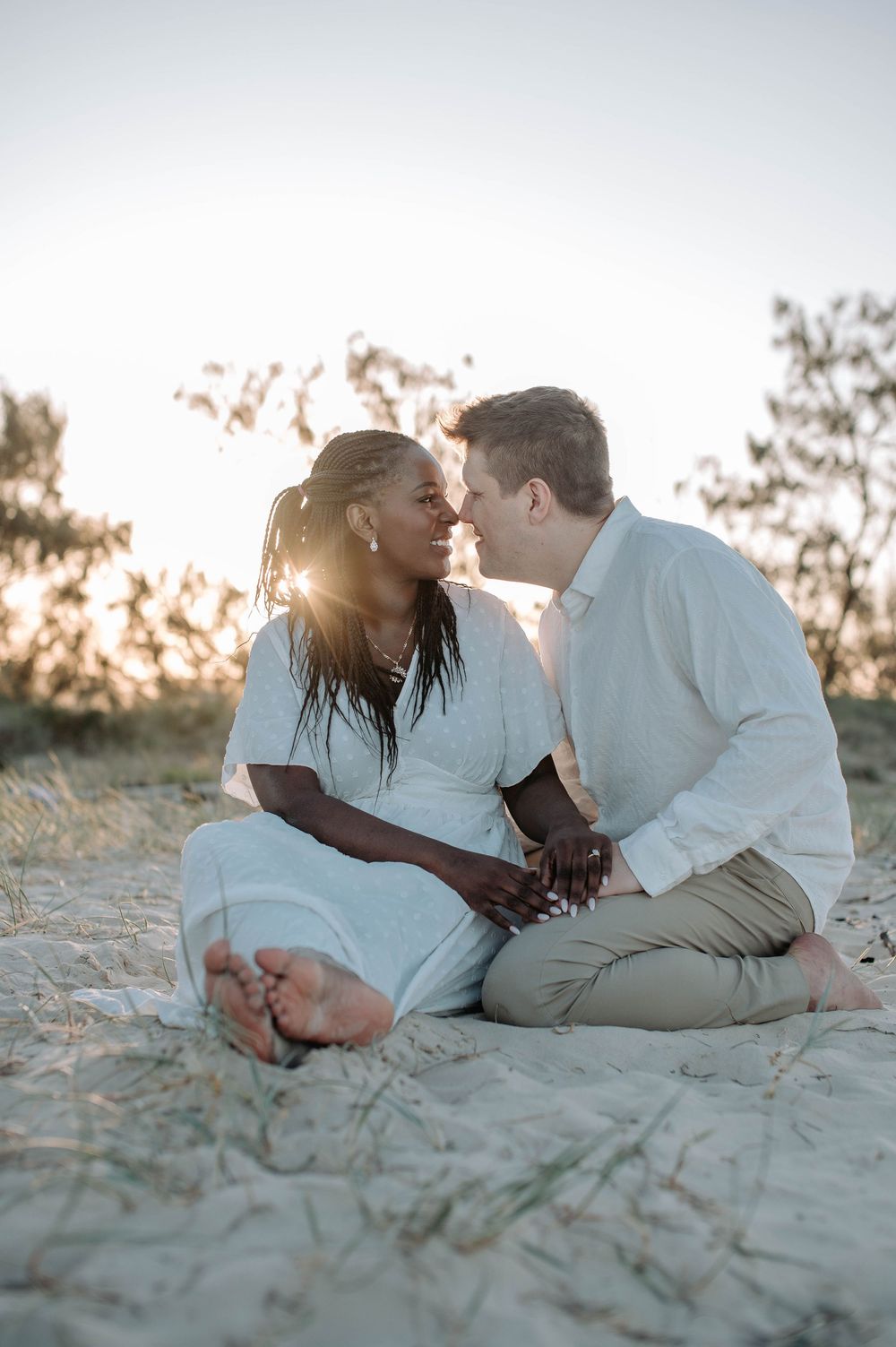 A romantic couple laying together on sandy beach dunes during sunset, dressed in white casual attire.