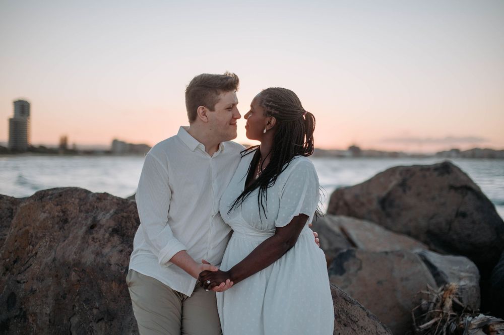 A romantic couple embraces on rocky shoreline during sunset with cityscape in background.