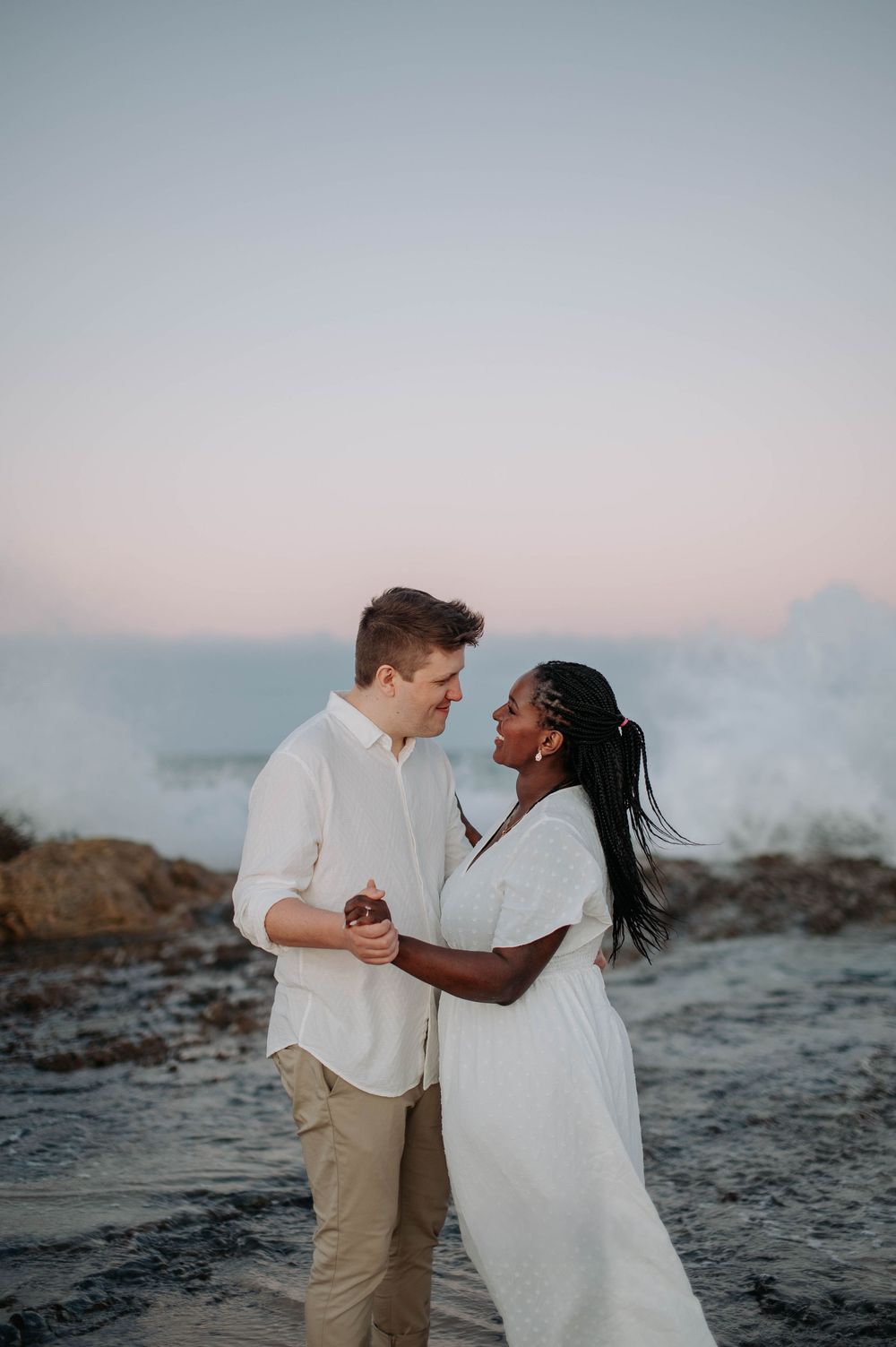 A romantic moment shared between a couple on a rocky beach at dusk with misty waves in the background.