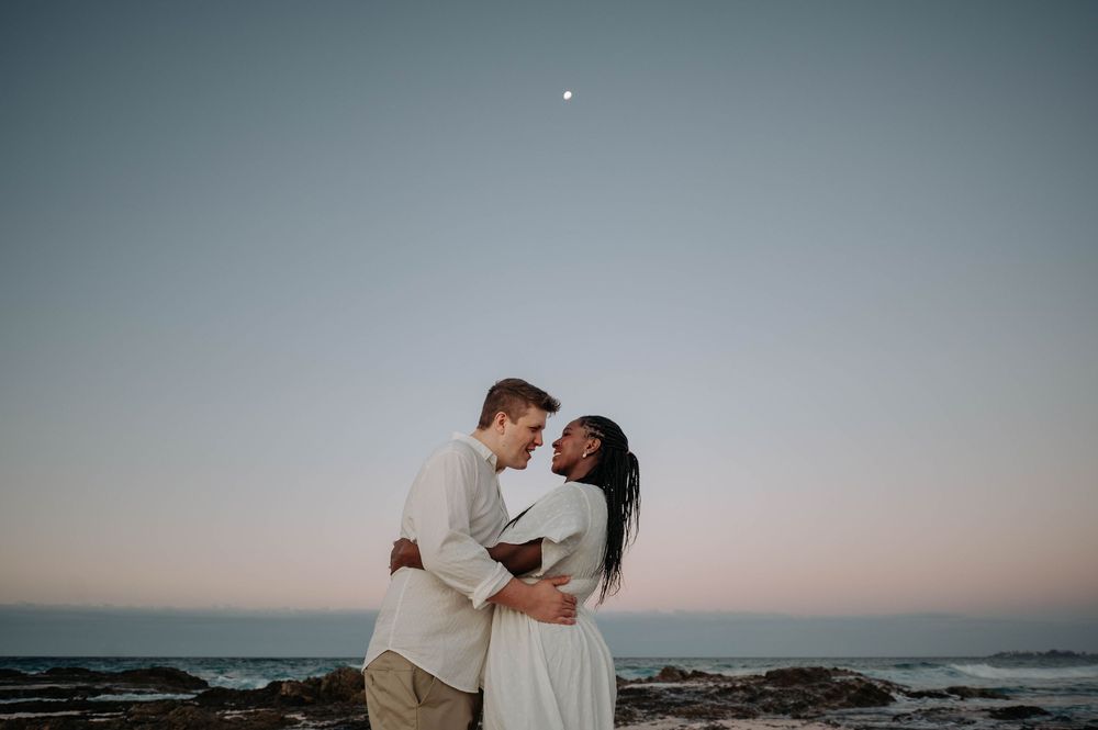 Couple embracing against a dusky ocean backdrop with a small moon visible in the twilight sky.
