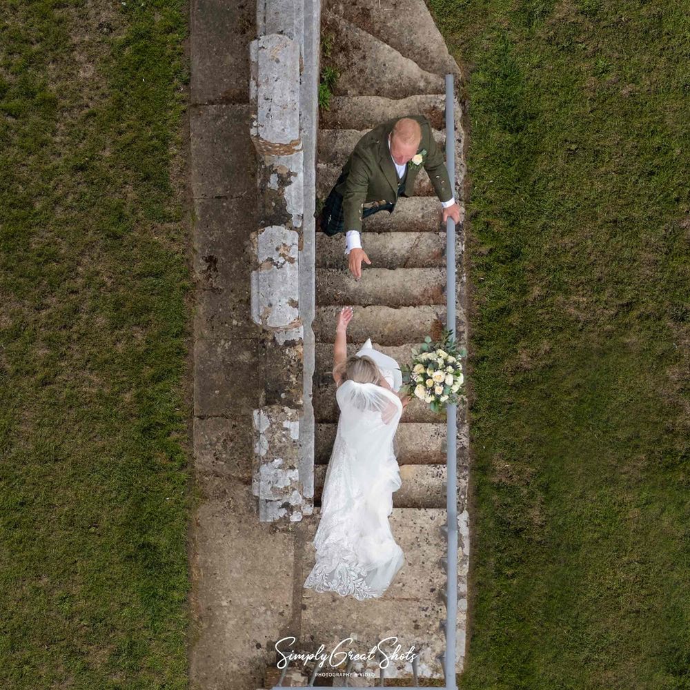 Fort Henry wedding photographer caputres with a drone the groom offering his hand to his bride as she walks down an old exterior staircase.