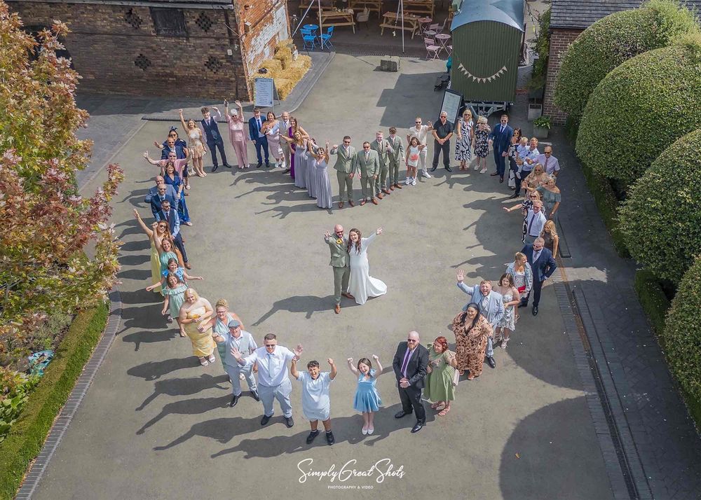 The wedding party form a heart shape with the bride and groom in the middle which is captured with a drone from from above.