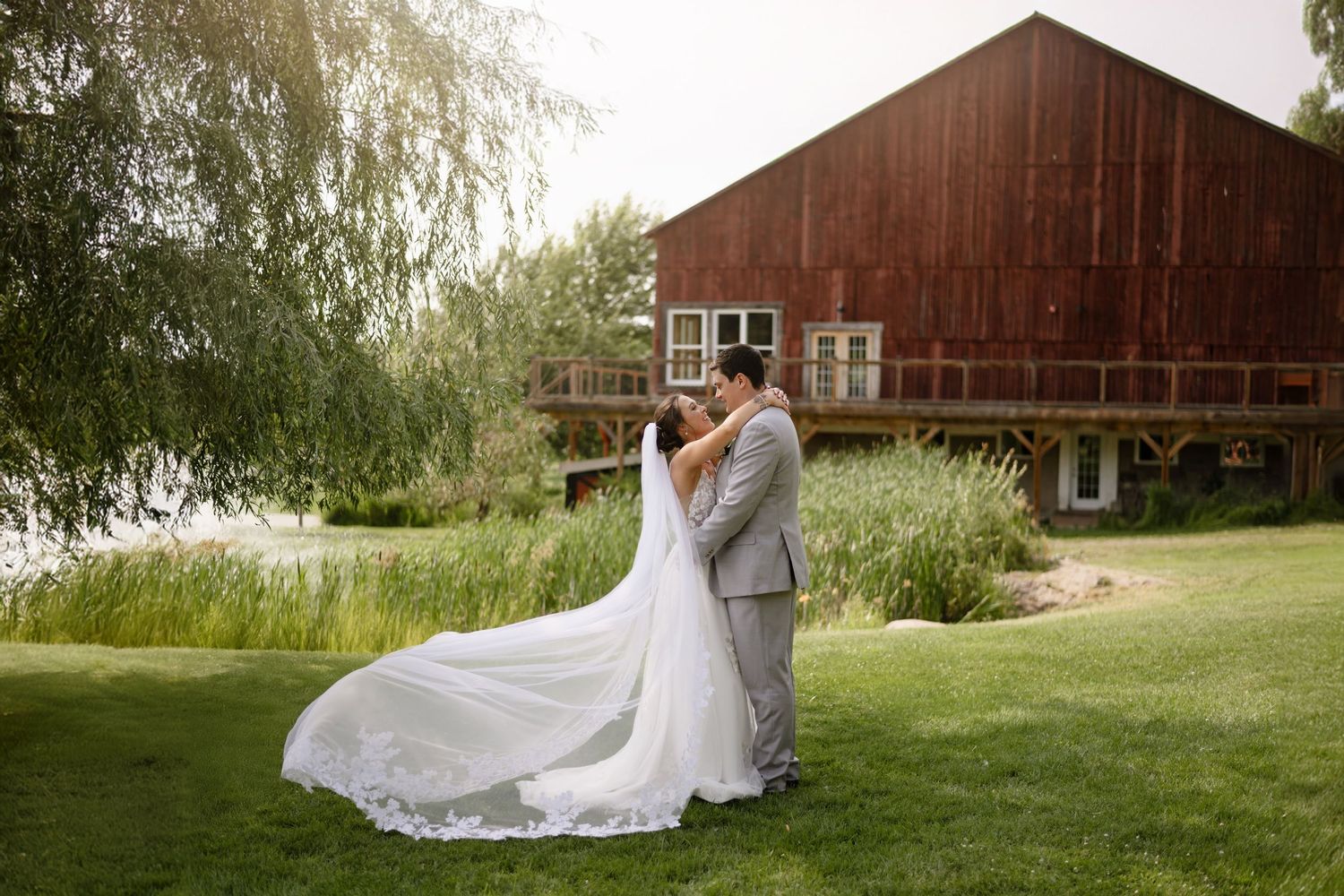 A couple embraces on a manicured lawn in front of a rustic brown barn with a white wedding dress trailing behind.