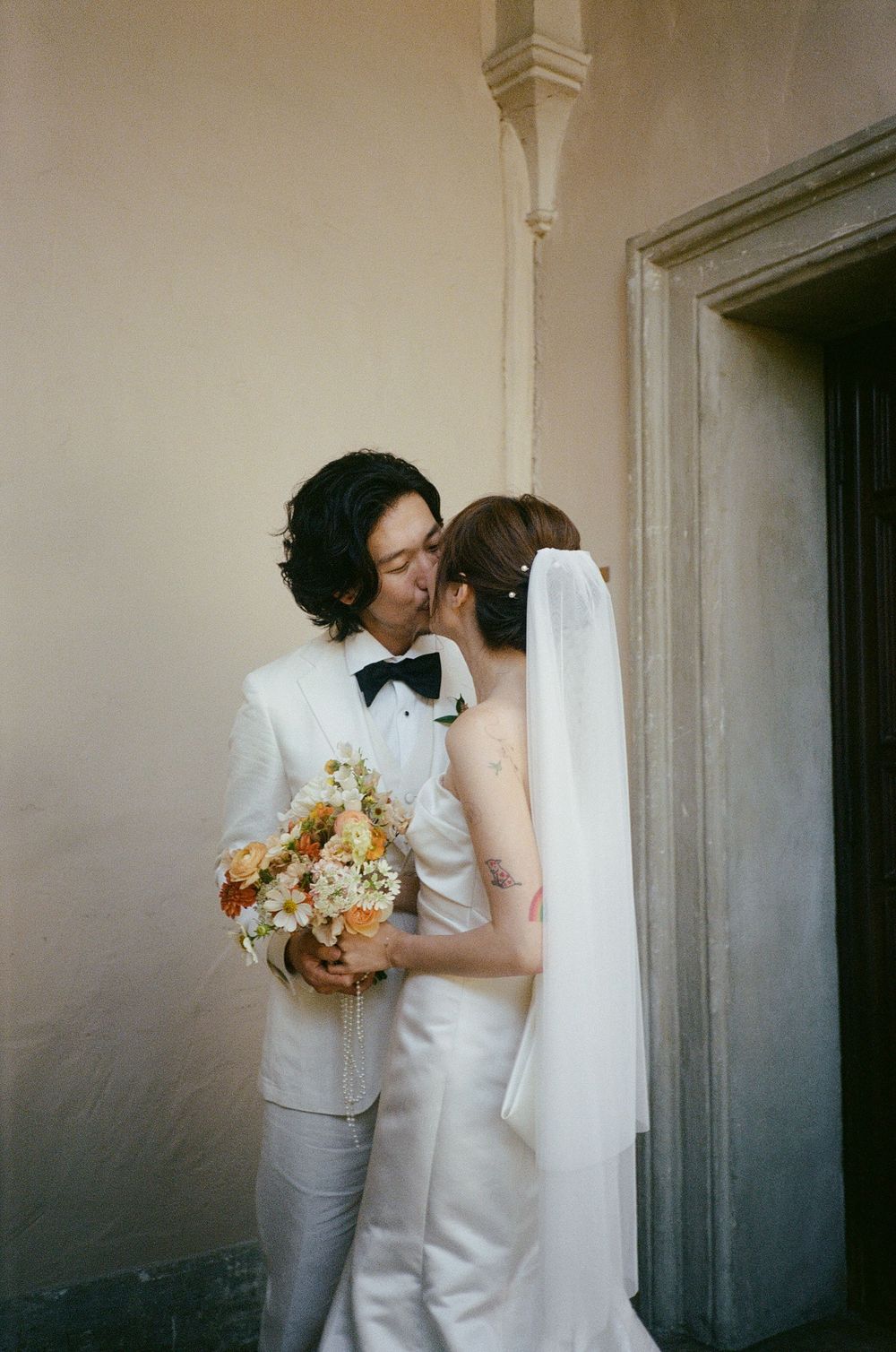 Romantic wedding portrait of a couple in white with peach-toned bouquet by an ornate doorway.