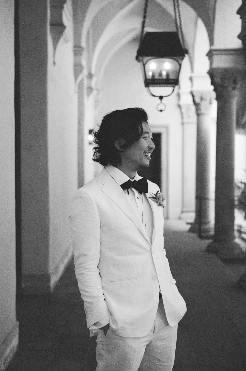 A person in a white tuxedo and bowtie stands under an elegant archway with hanging lanterns.