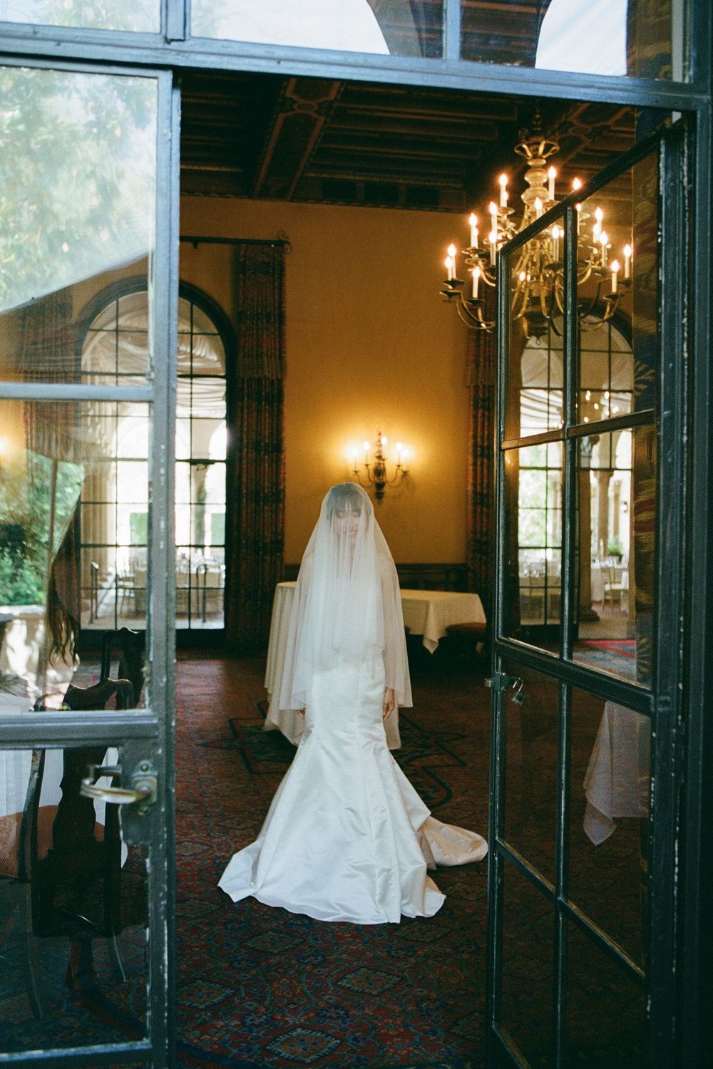 A bride in a flowing white wedding gown stands in an elegant room with arched windows and chandeliers.