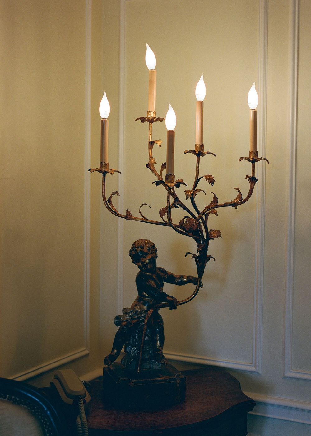 Ornate antique candelabra with cherub figure base holding multiple lit candles against beige wall.