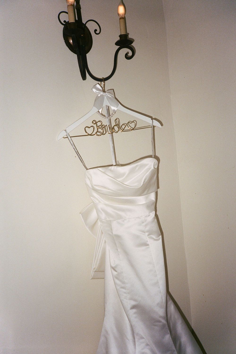 An elegant white wedding dress hangs on a decorative hanger beneath a wall-mounted candle sconce.