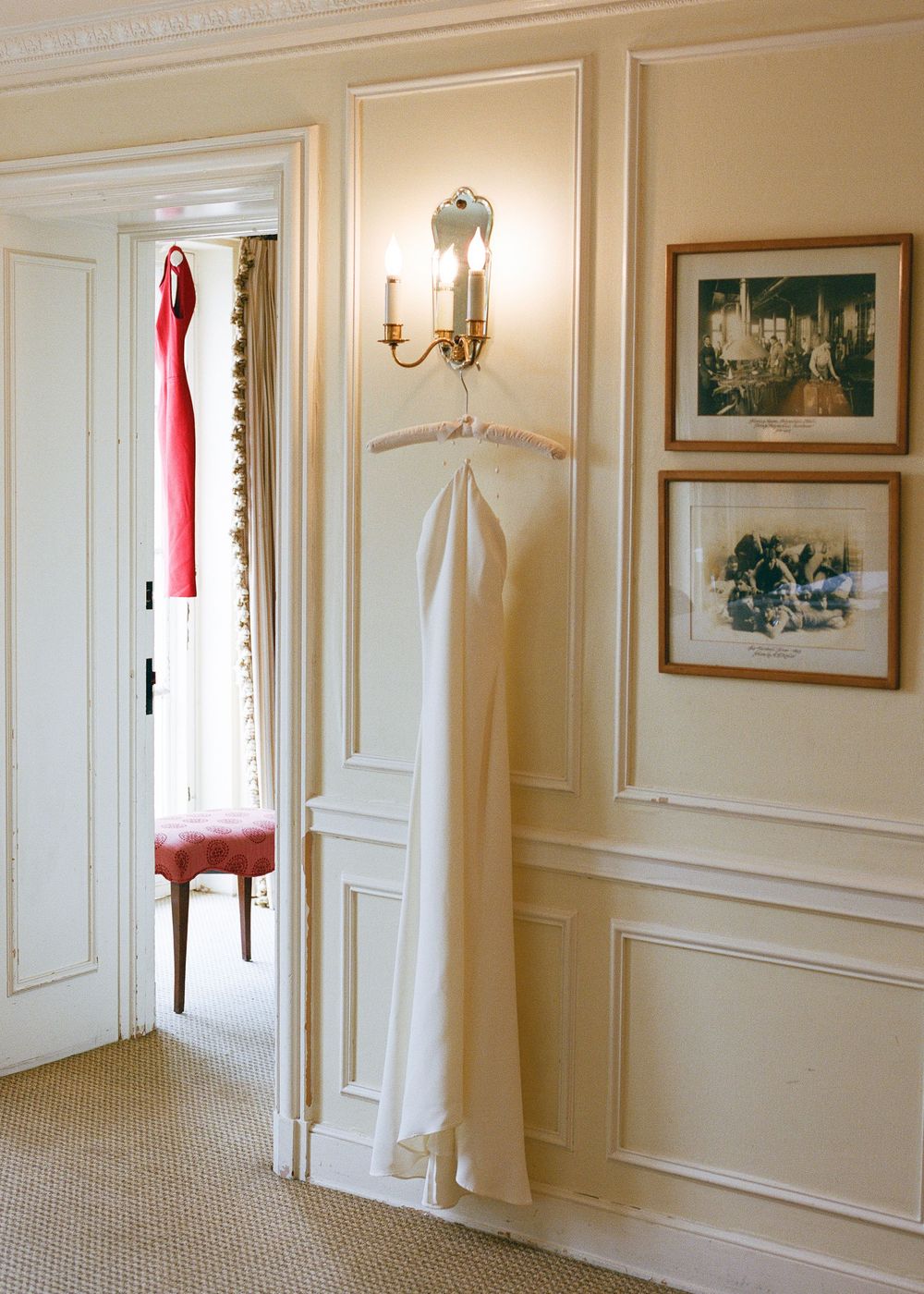 Wedding dress and red gown hanging in an elegant room with wainscoting and framed artwork.