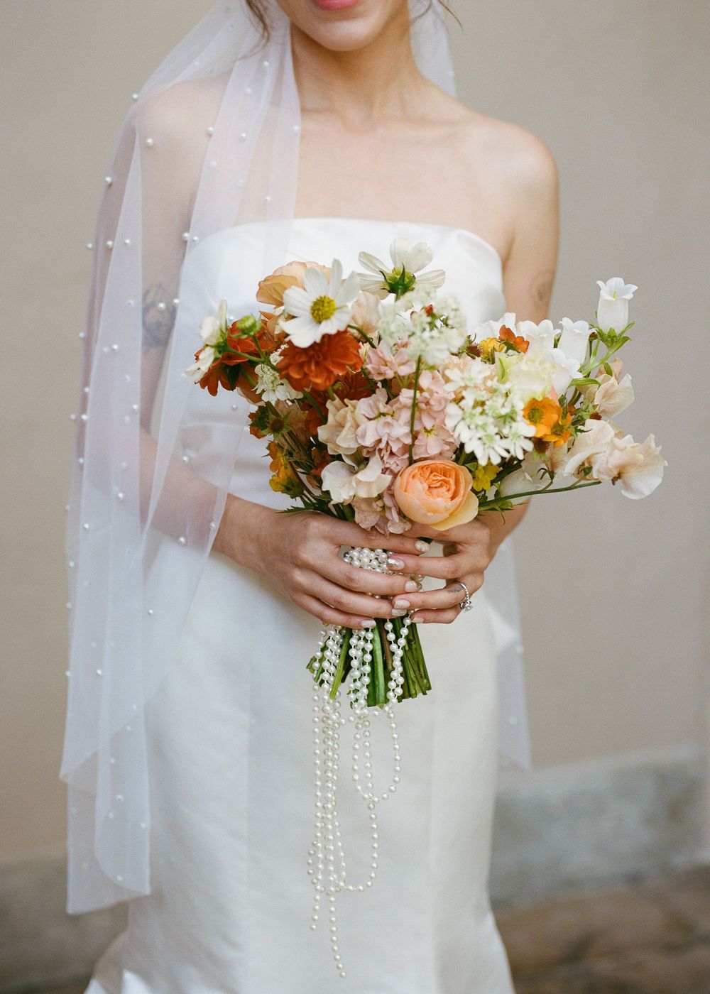 Bridal bouquet of mixed flowers with pearls held against white wedding dress.