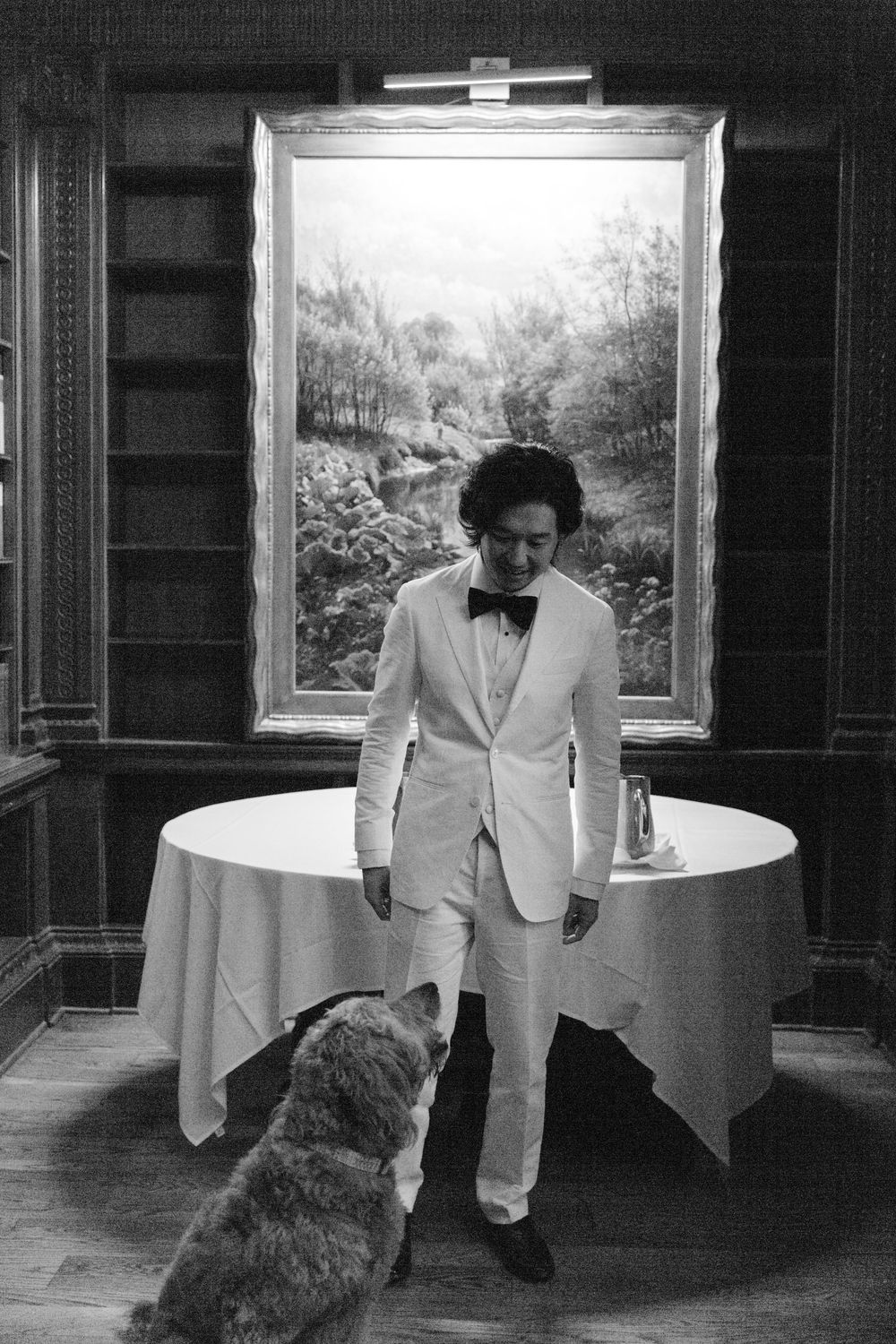 A person in a white tuxedo stands by a round table while greeting a dog in an elegant room.