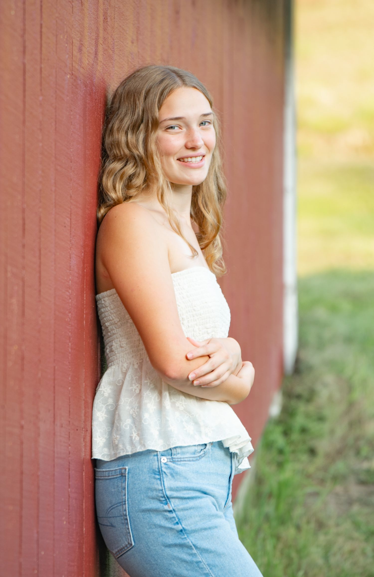 Portrait series of someone in a white sleeveless top and blue jeans posing against a red barn wall in natural light.
