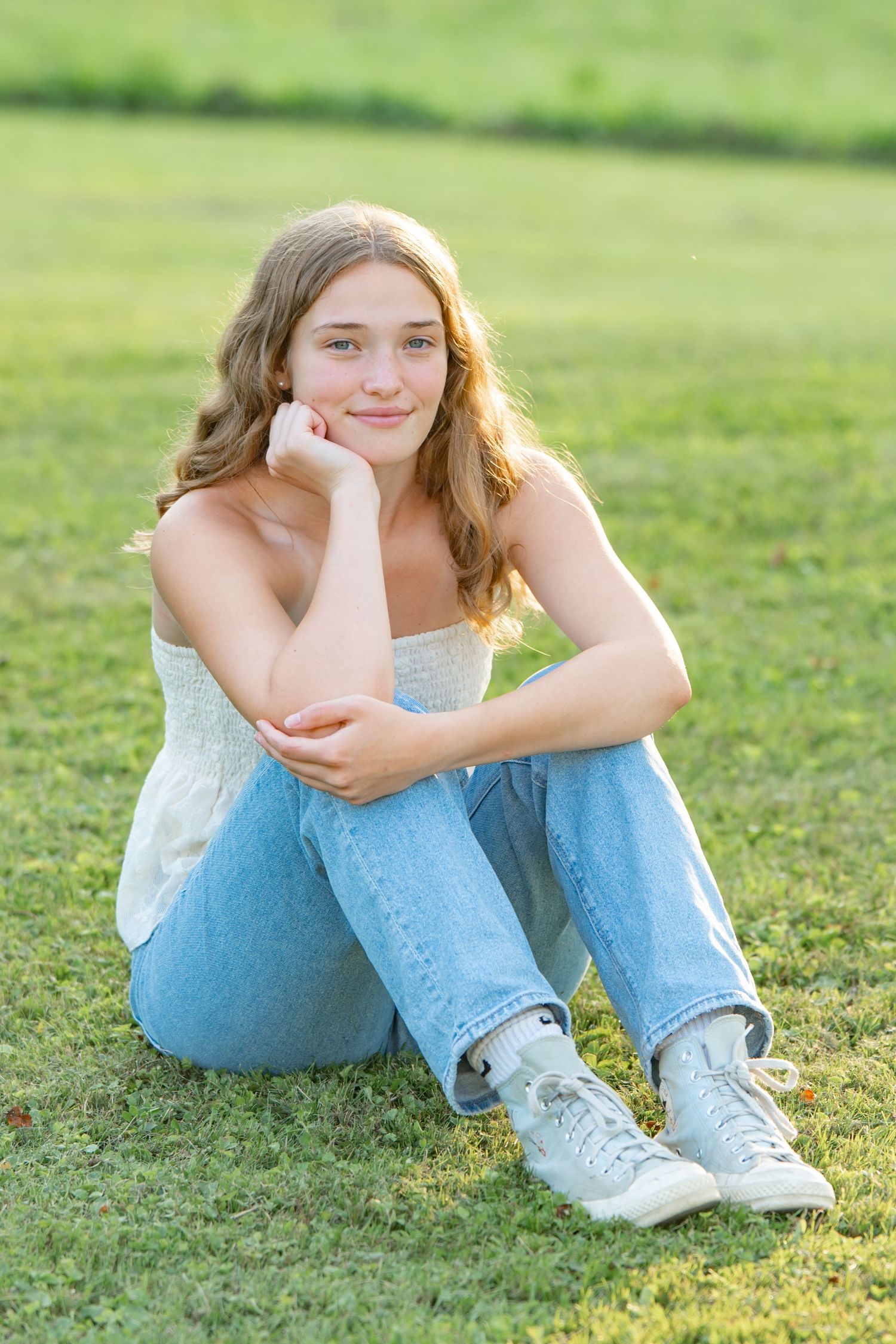Casual summer portrait sitting on grass wearing white top, jeans and sneakers in golden sunlight.