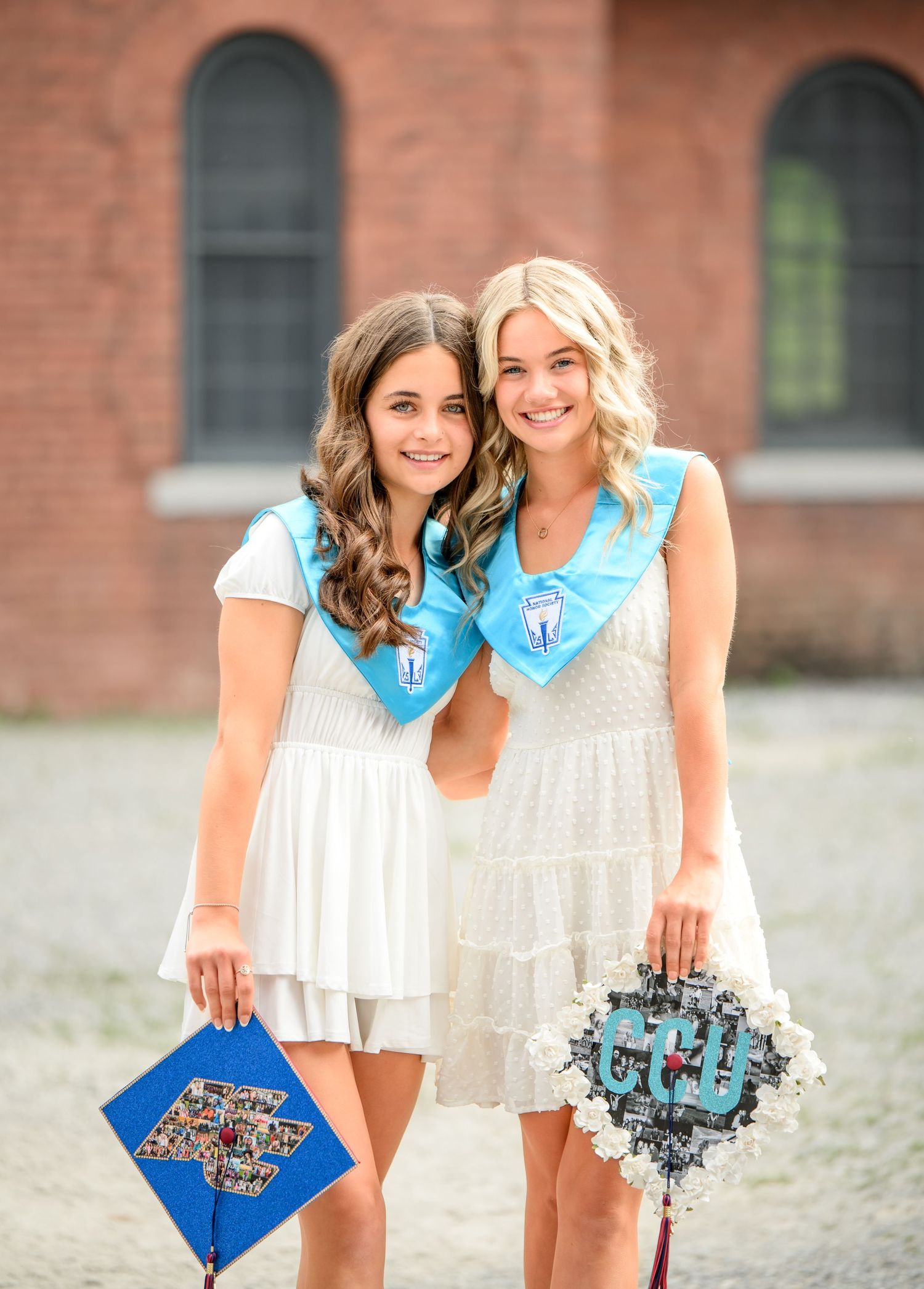 Two friends in matching white outfits holding floral decorative clutches pose against a brick building backdrop.