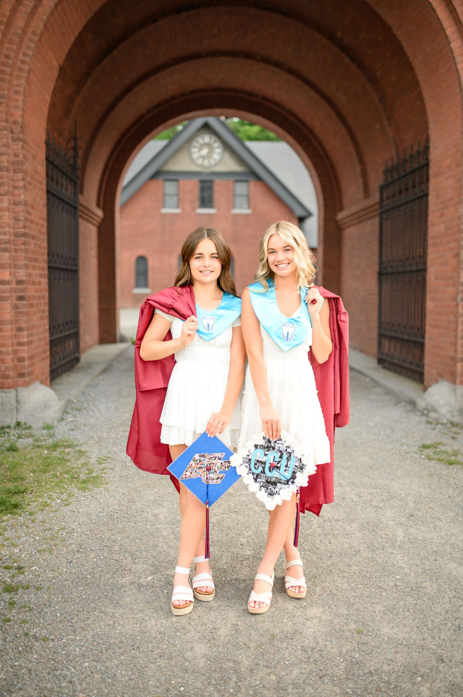 Two graduates in white dresses and red graduation gowns hold decorated caps while standing under a brick archway.