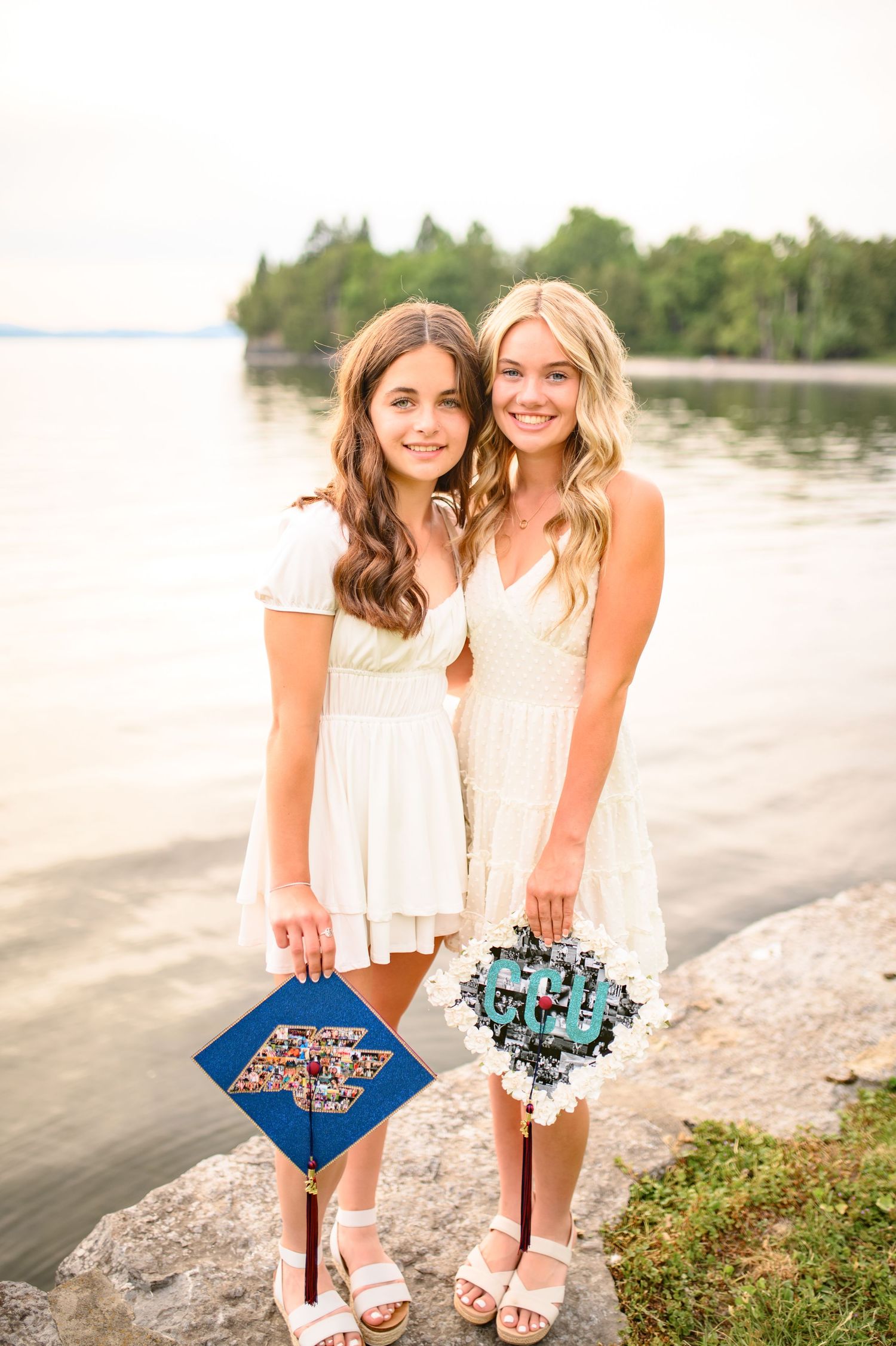Two friends in white summer dresses holding decorative clutches pose together by a lakeside.