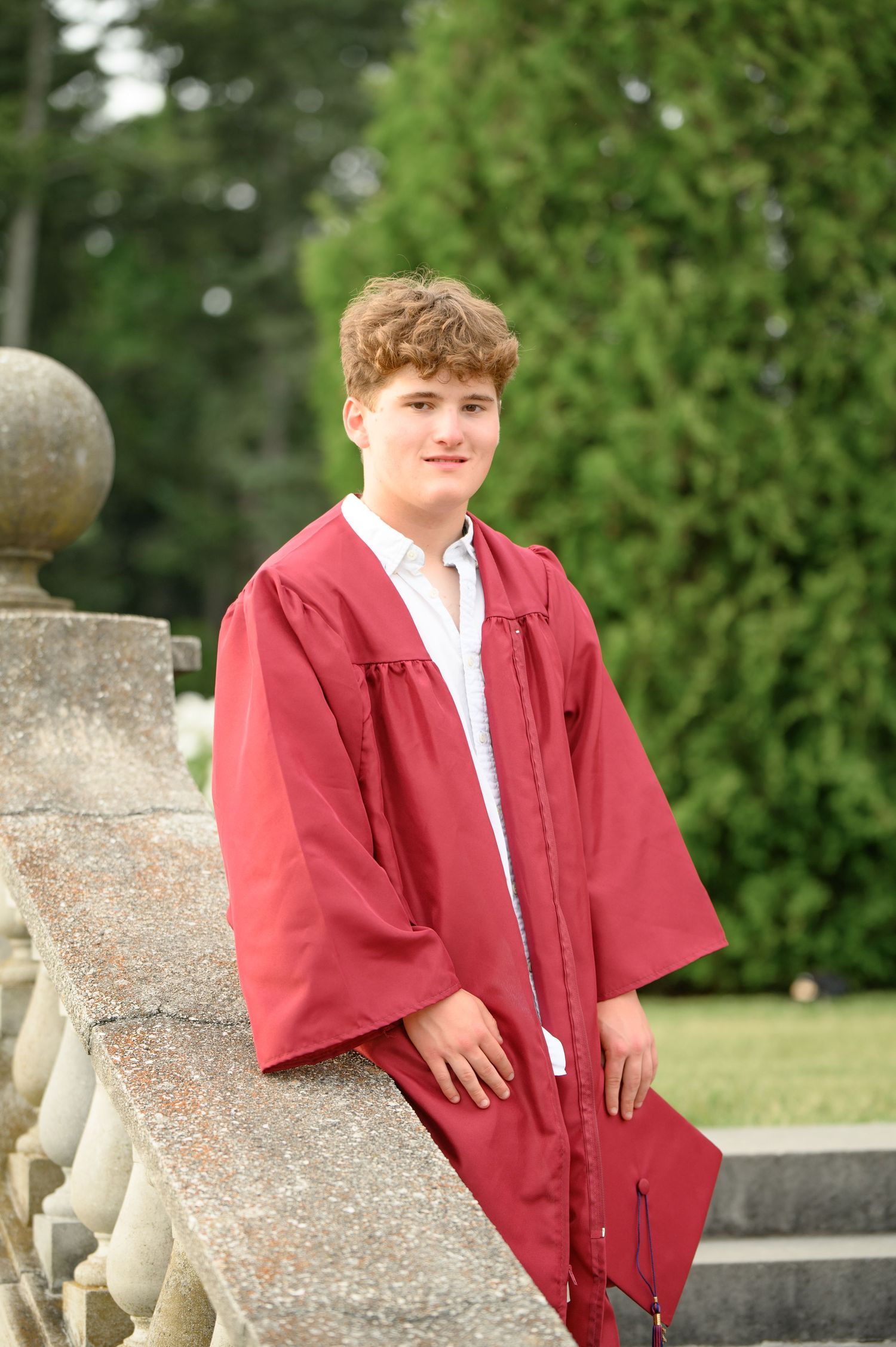 Graduate in burgundy academic regalia stands against stone railing and greenery in outdoor setting.