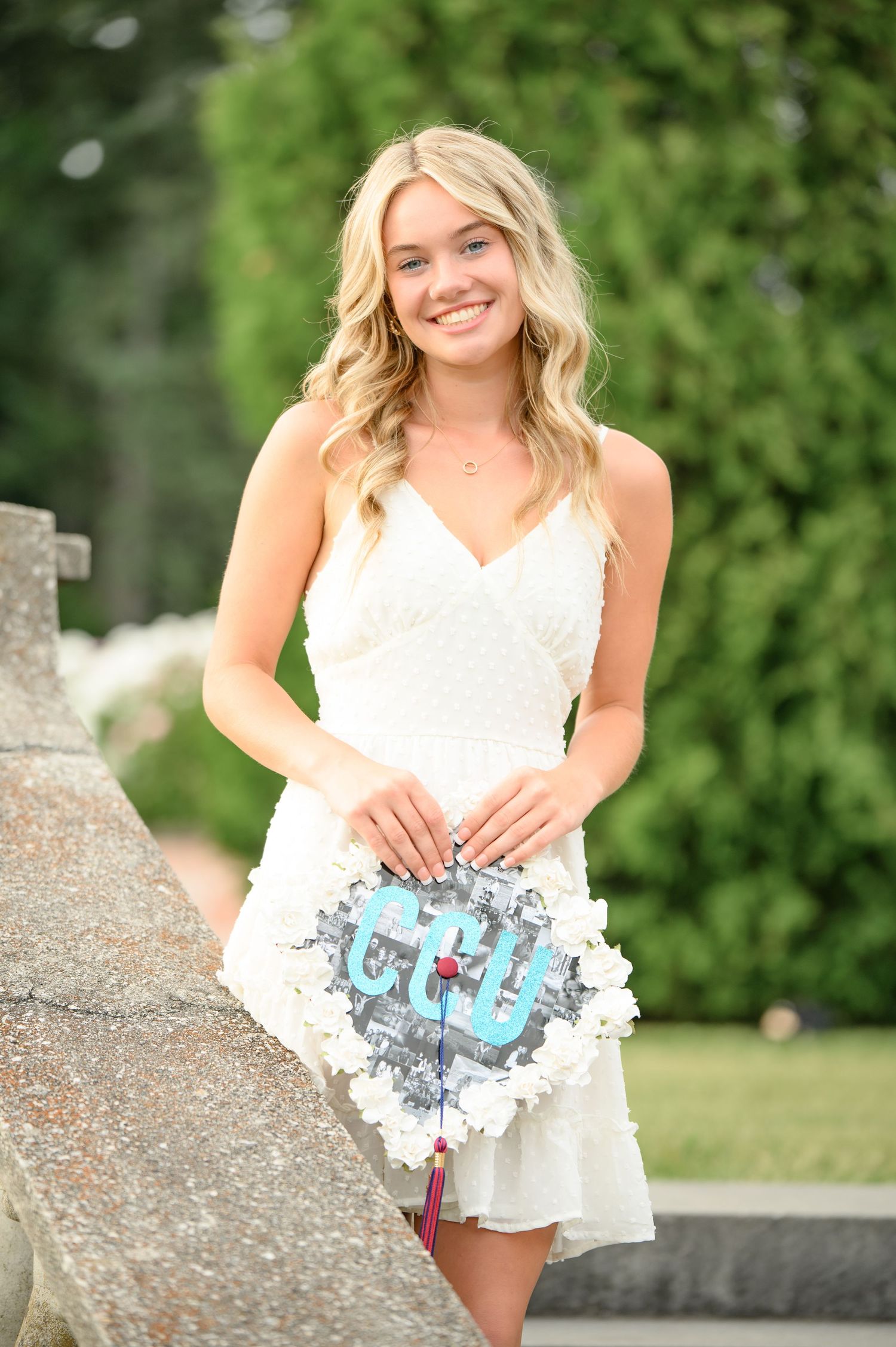 Graduate in white lace dress poses outdoors while holding decorated graduation cap with blue monogram design.