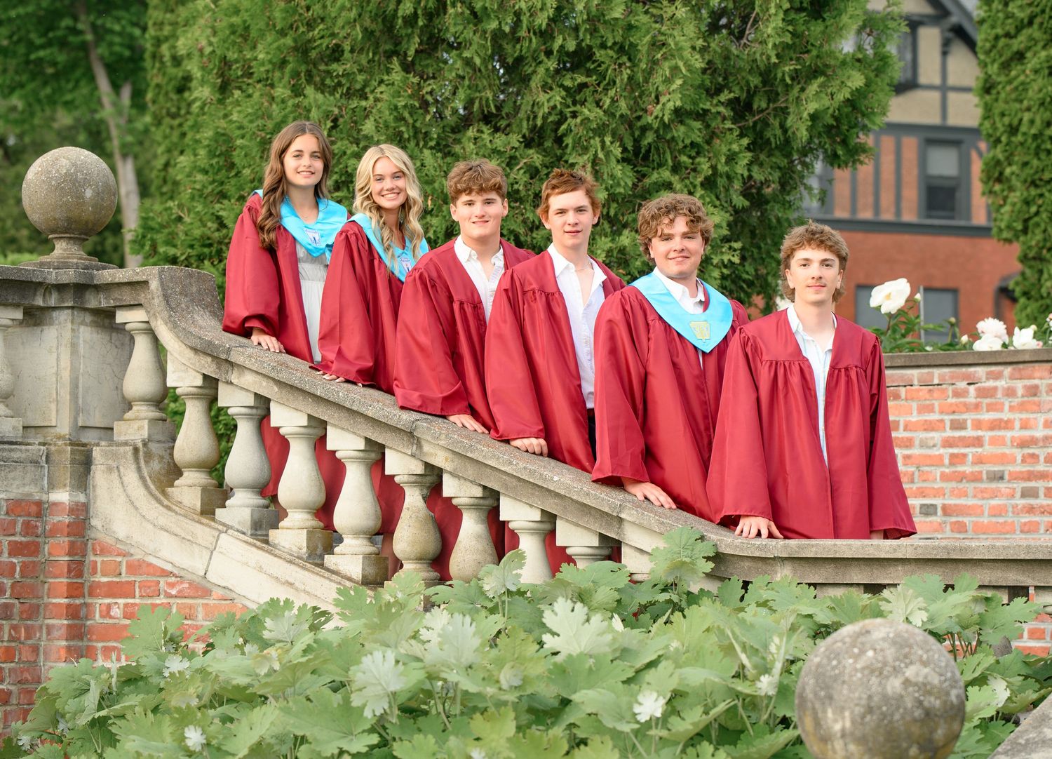 Choir members in red robes pose on outdoor stone stairs surrounded by greenery.