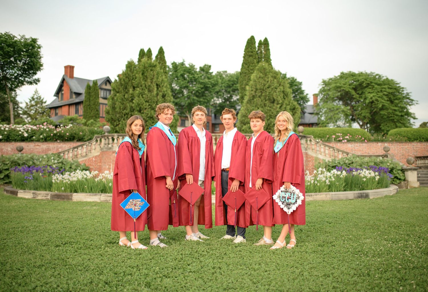 A line of graduates in red caps and gowns standing on grass with trees in the background.