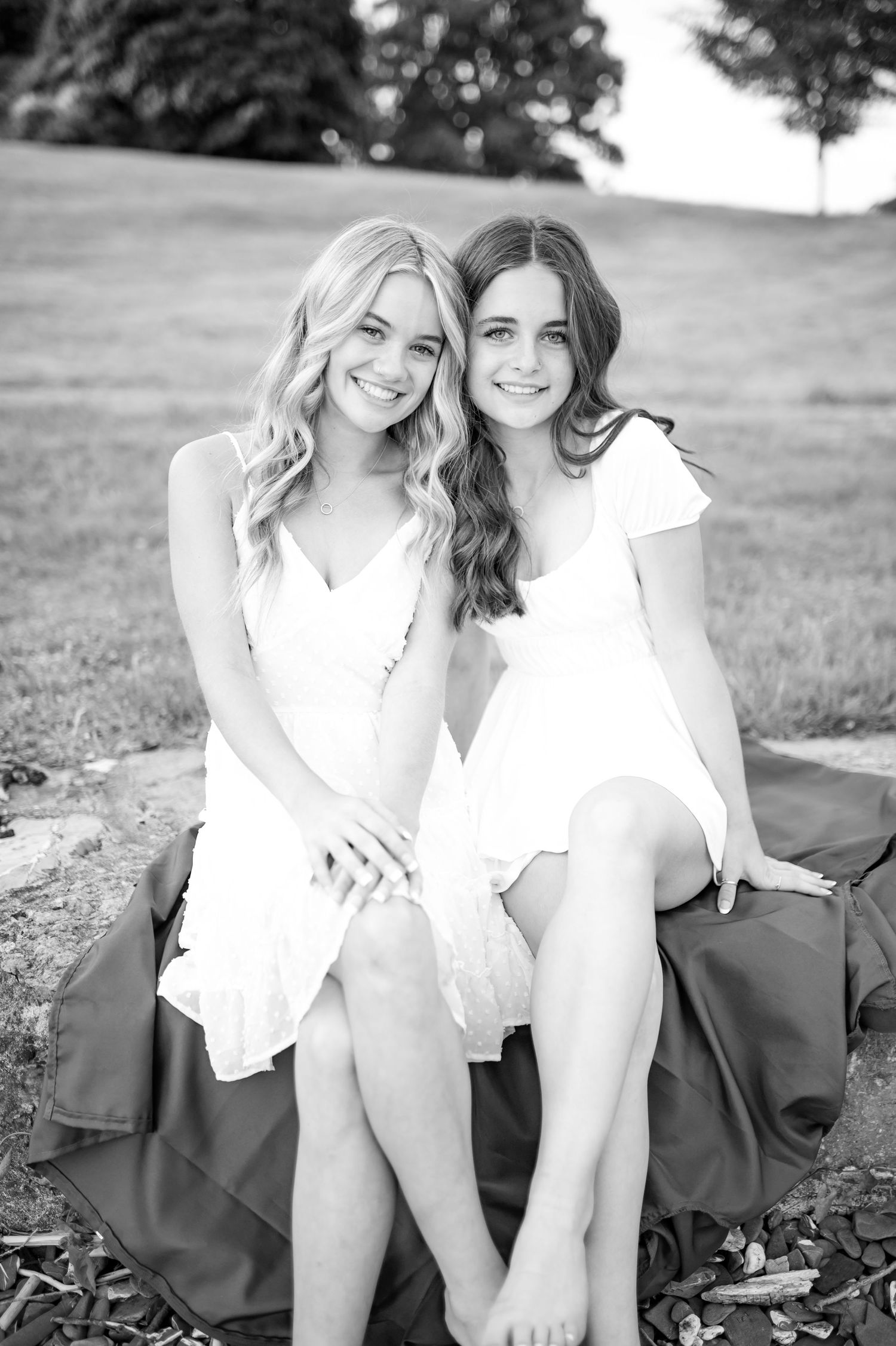 Two friends in white summer dresses sitting together outdoors in black and white photo.