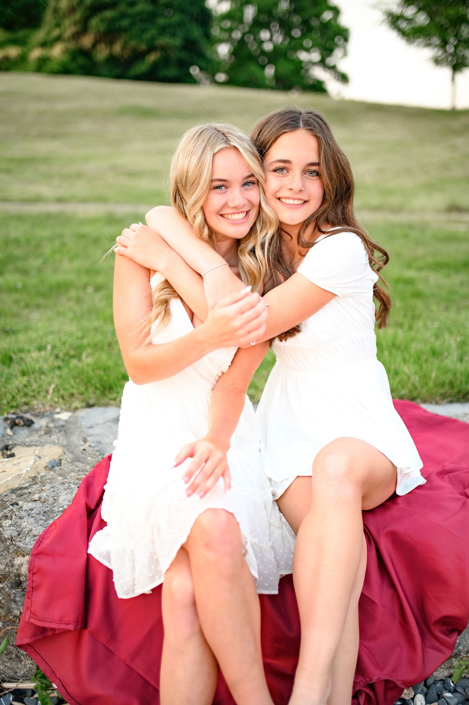 Two friends in white dresses share a candid moment while sitting on a burgundy blanket outdoors.