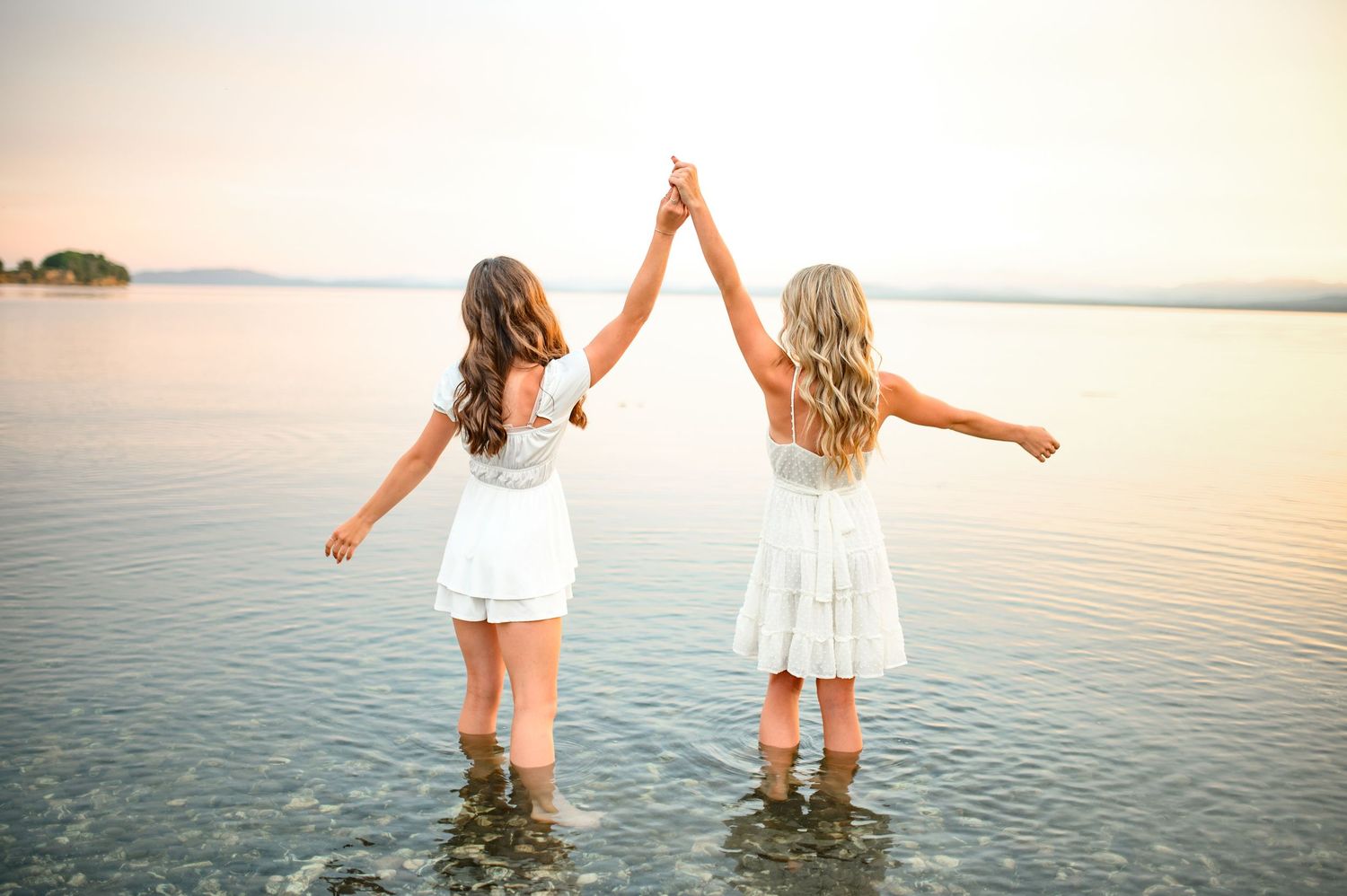 Two people in white dresses hold hands while standing in calm water at sunset by the ocean.